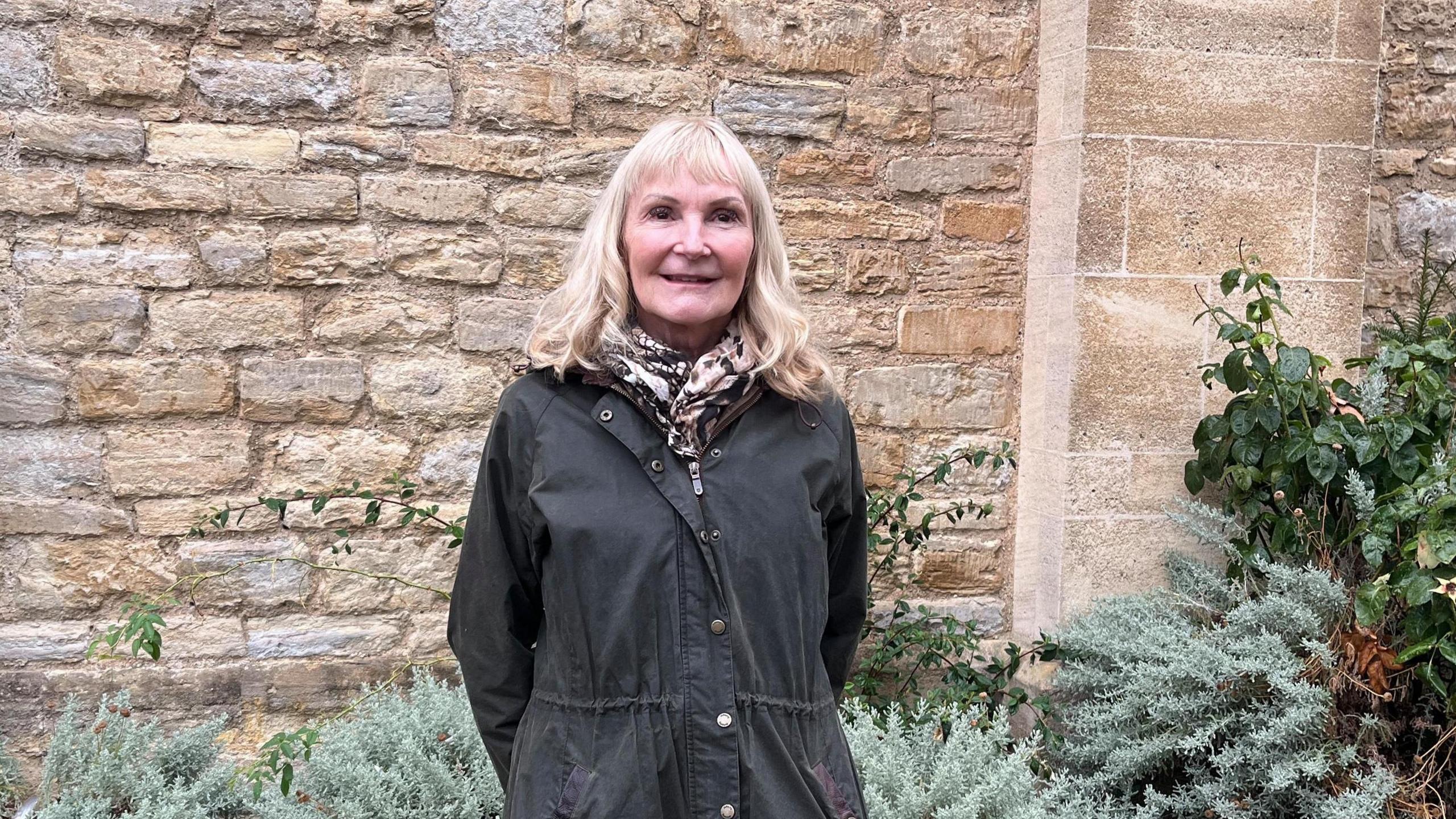 A woman with long grey hair wearing a scarf and a green coat standing in front of a church wall. There are bushes by the wall.