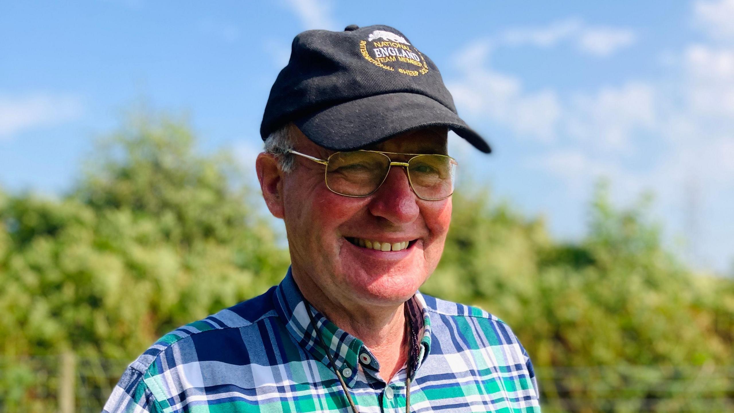 Farmer Philip Walton standing in the field. He is wearing a black hat, glasses and blue, white and green checkered shirt. He is smiling in the sunshine.  