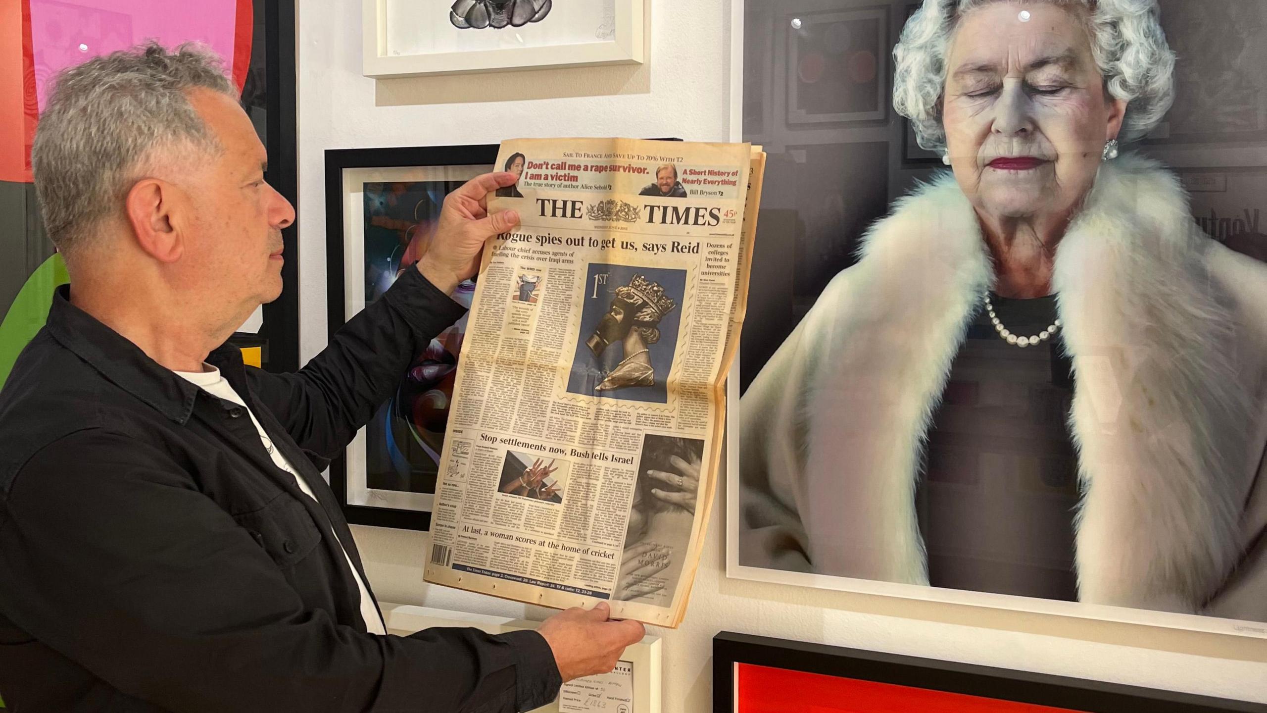 Enter Gallery founder Lawrence Alkin holding a newspaper in front of a wall full of artwork. 