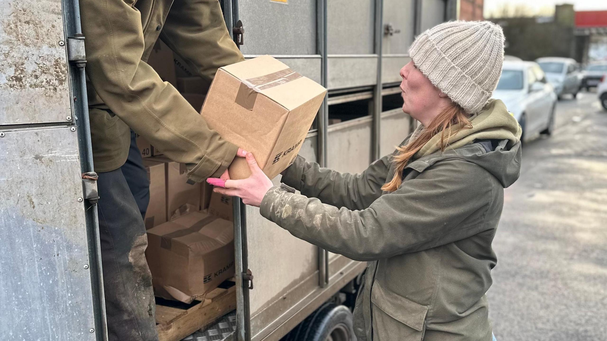 Farmer Charlotte Ashley, who wears a khaki wax coat and a cream bobble hat, lifts a box passed to her from a sheep truck