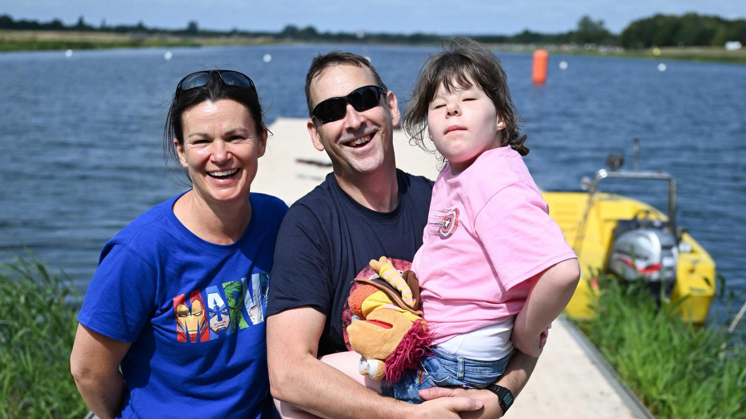 A woman wearing a blue t-shirt, standing next to a man wearing sunglasses who is holding a young girl, wearing a pink t-shirt. There is a lake behind them.