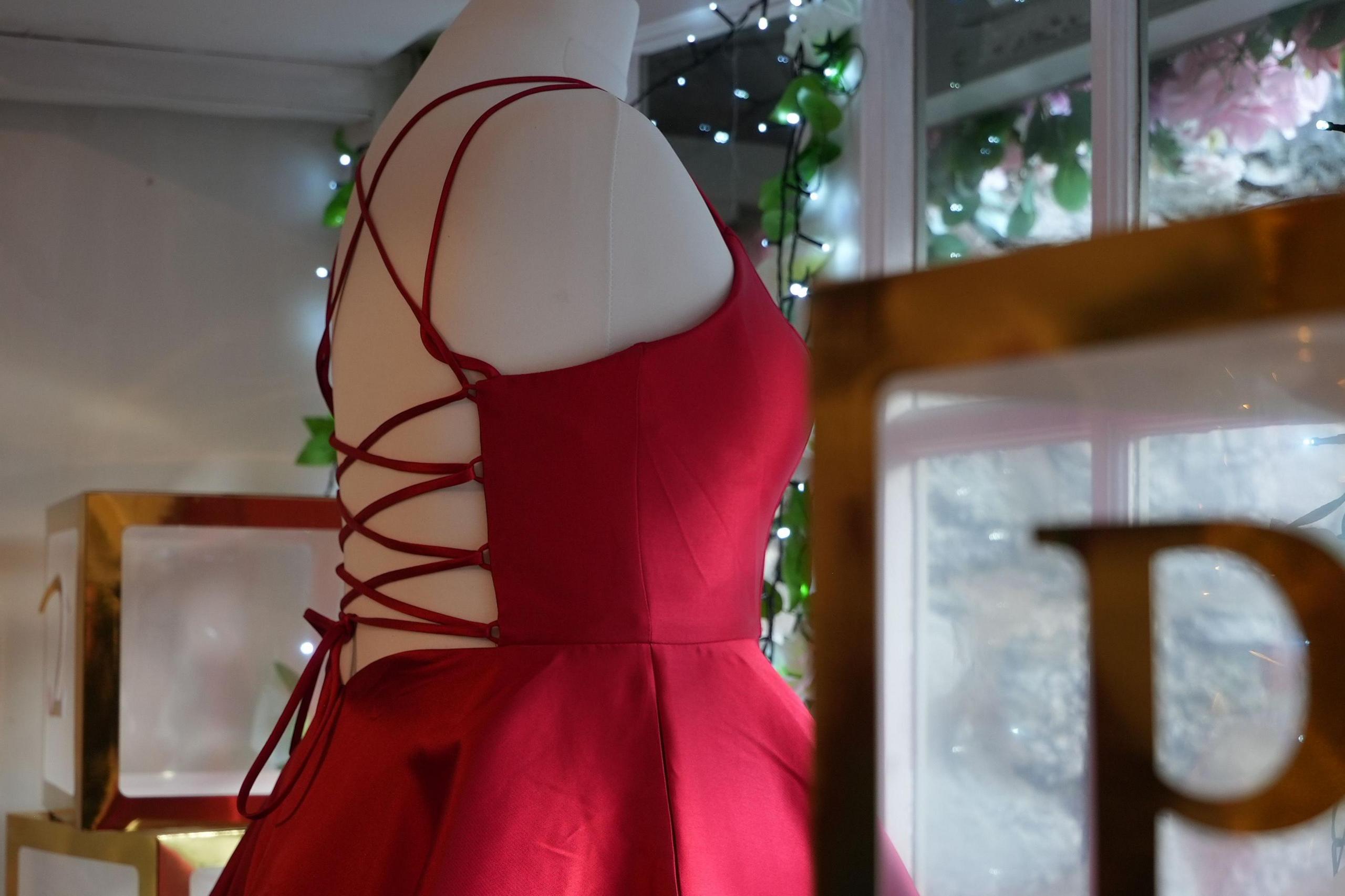 A red satin dress on a mannequin inside a shop window. The picture is taken from within the shop. The dress has thin straps and a lace-up back.