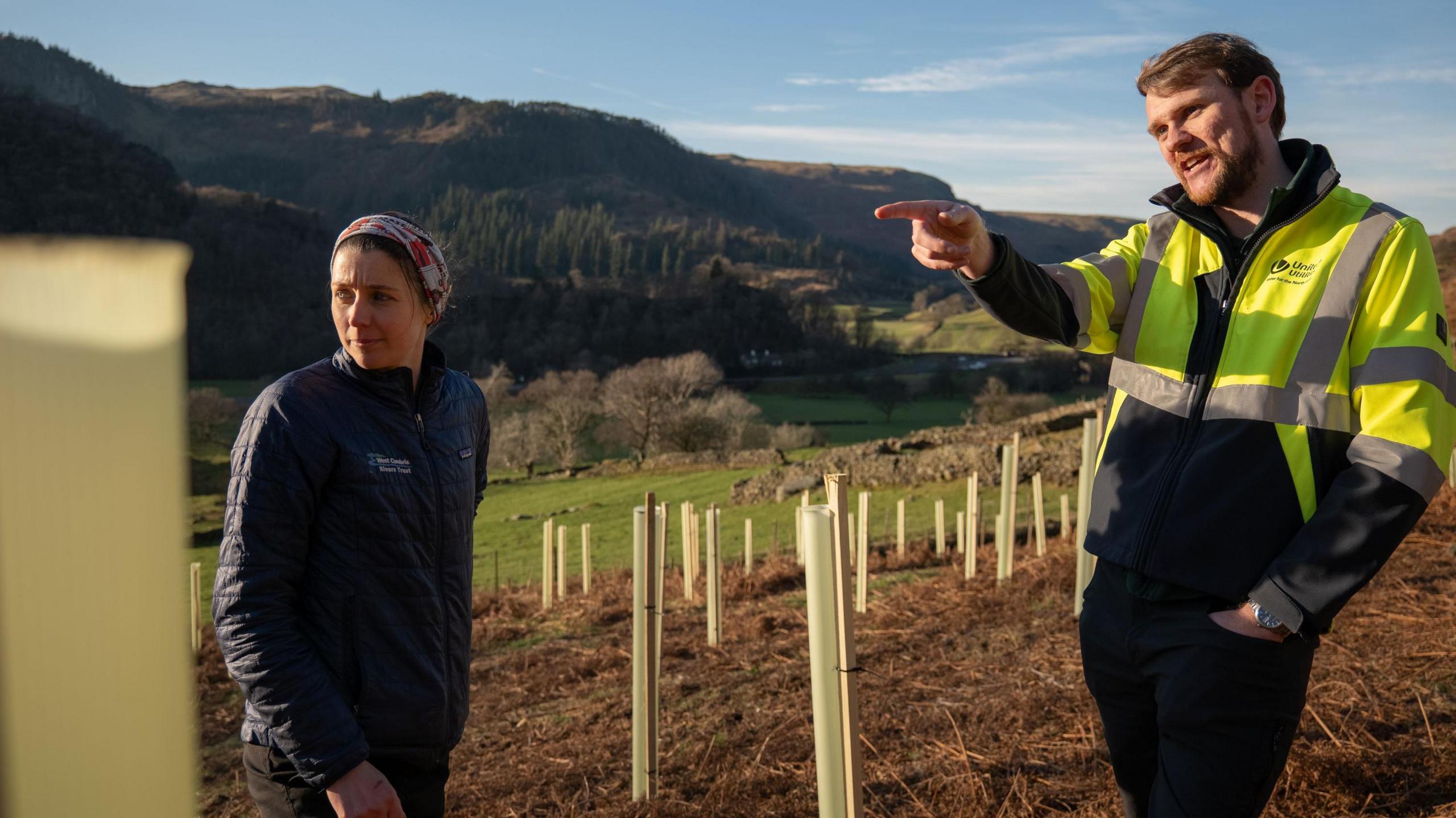 West Cumbria Rivers Trust's project manager, Caitlin Pearson, wears dark clothing and stands talking to Andrew Wright, who is wearing a fluorescent United Utilities jacket. They are standing where the trees have been planted.