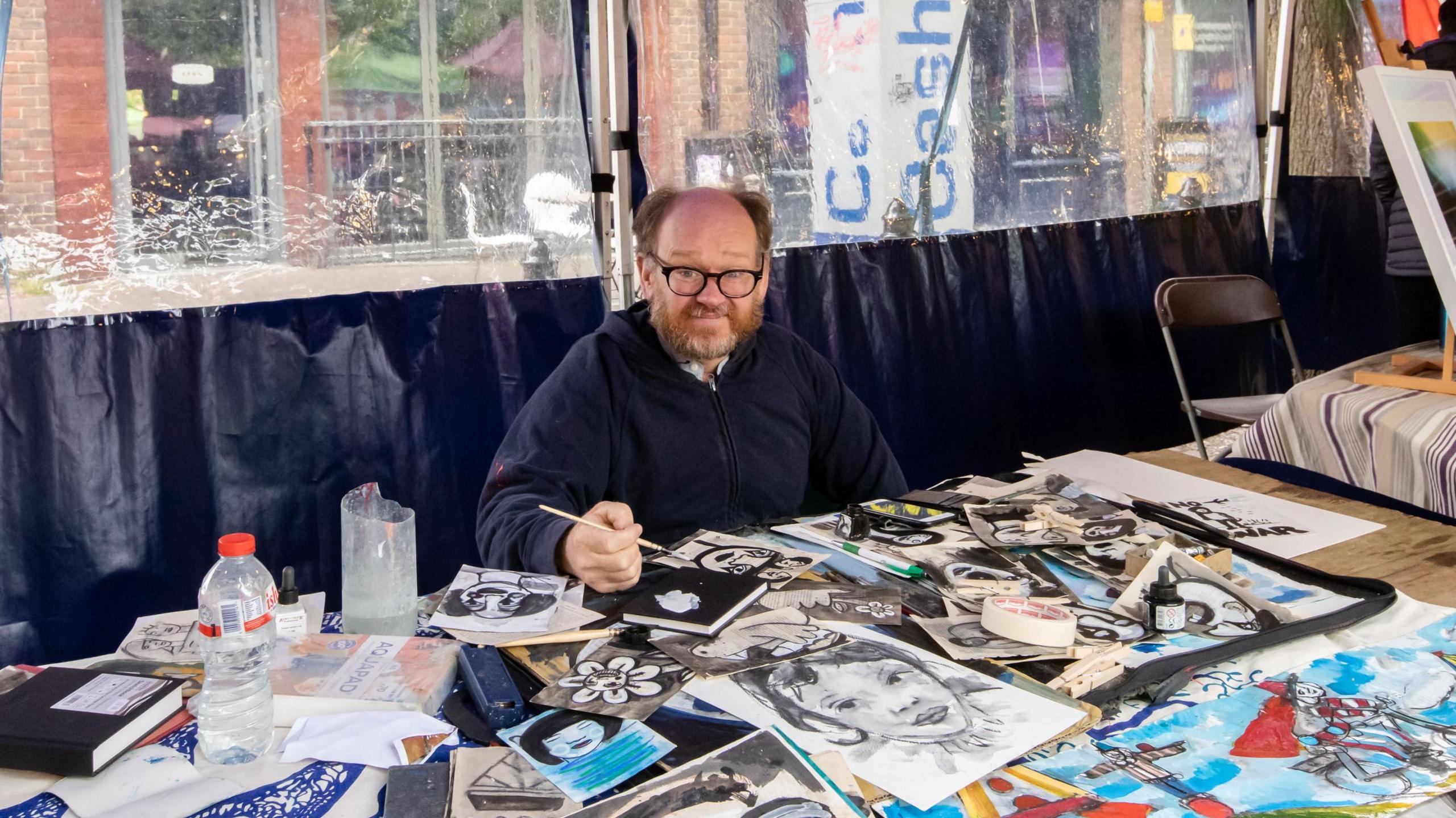 A man with a beard and glasses sits smiling at a stall holding a paintbrush, with sketches and paintings displayed in front of him