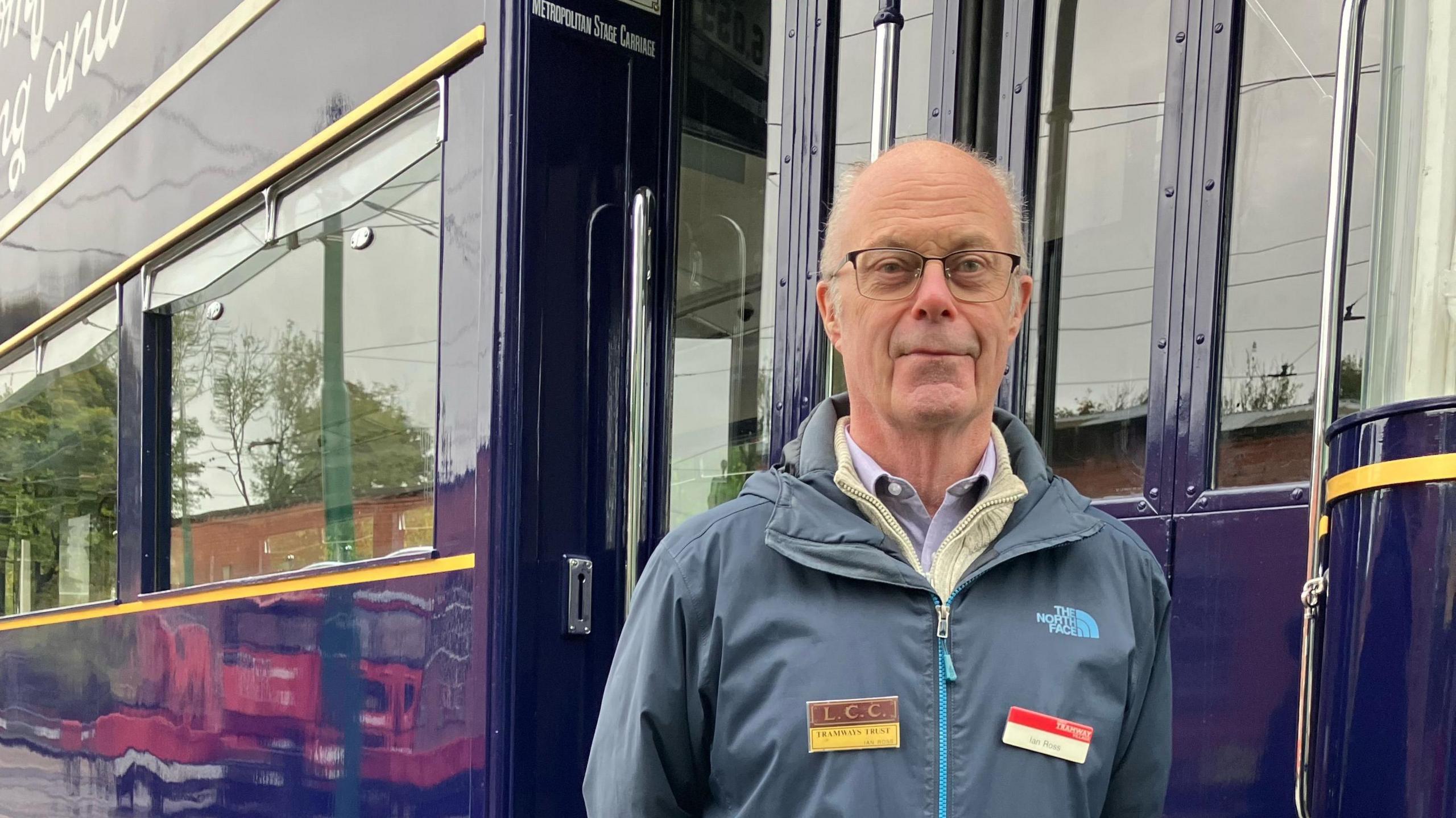 A gentleman with white hair stands in front of a blue tram car. He is wearing a navy zipped coat and glasses.