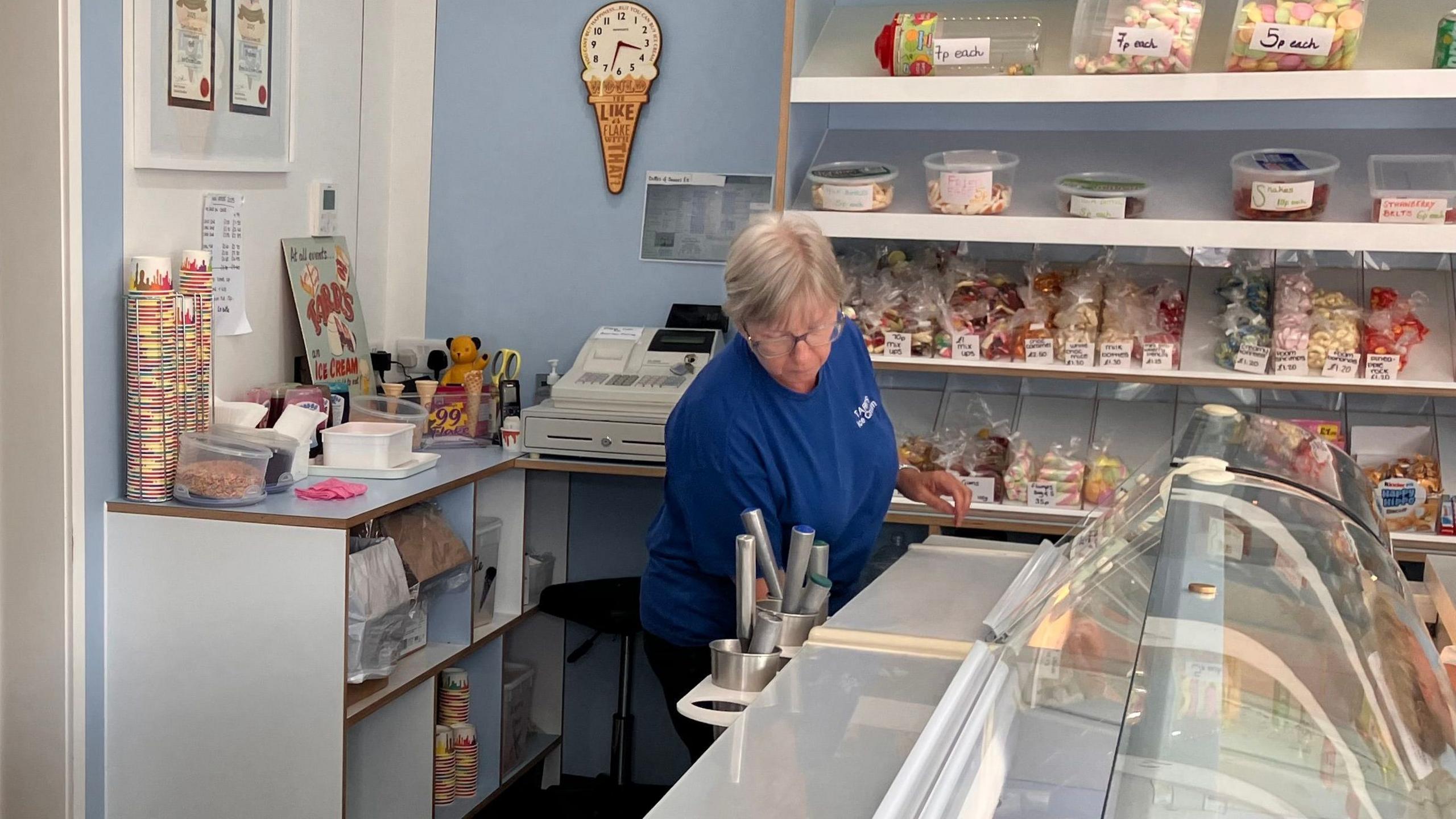 A family member in blue overalls behind the counter in the shop with sweets in clear plastic bags on the shelf behind her