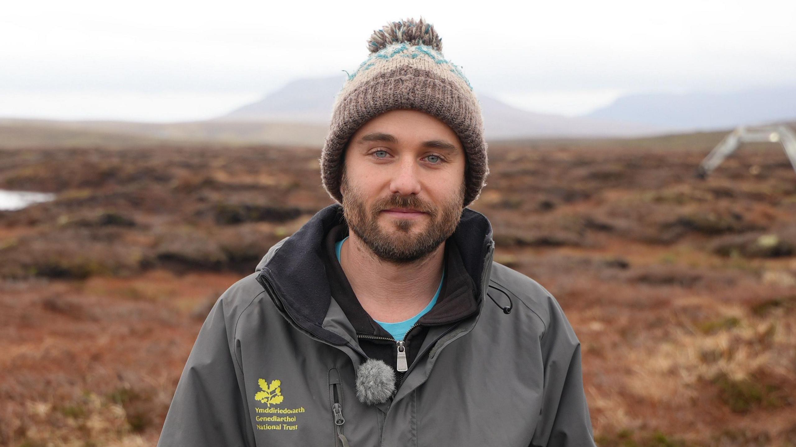 Iago Thomas, Swyddog Mawndiroedd yr Ymddiriedolaeth Genedlaethol, yn gwisgo het gaeaf a chot yr Ymddiriedolaeth Genedlaethol, gyda thir y Migneint yn y cefndir.