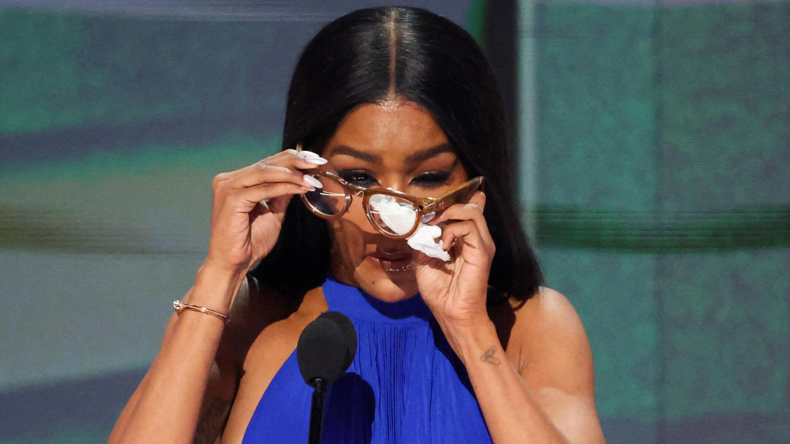 Teyana Taylor holding her glasses in front of her face and wiping the lenses with a tissue