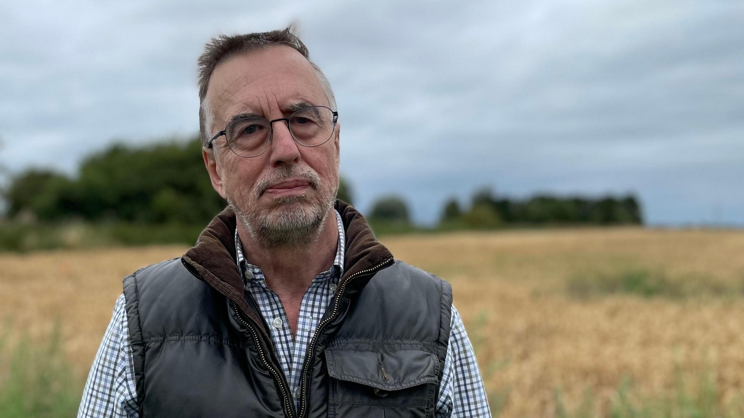 Stuart Gibbard, an agricultural historian, wears a serious expression as he stands in front of a field in the Lincolnshire Fens. He has short brown hair and a close-trimmed beard. He is dressed in a chequered shirt and dark gilet and wears glasses with thin frames 