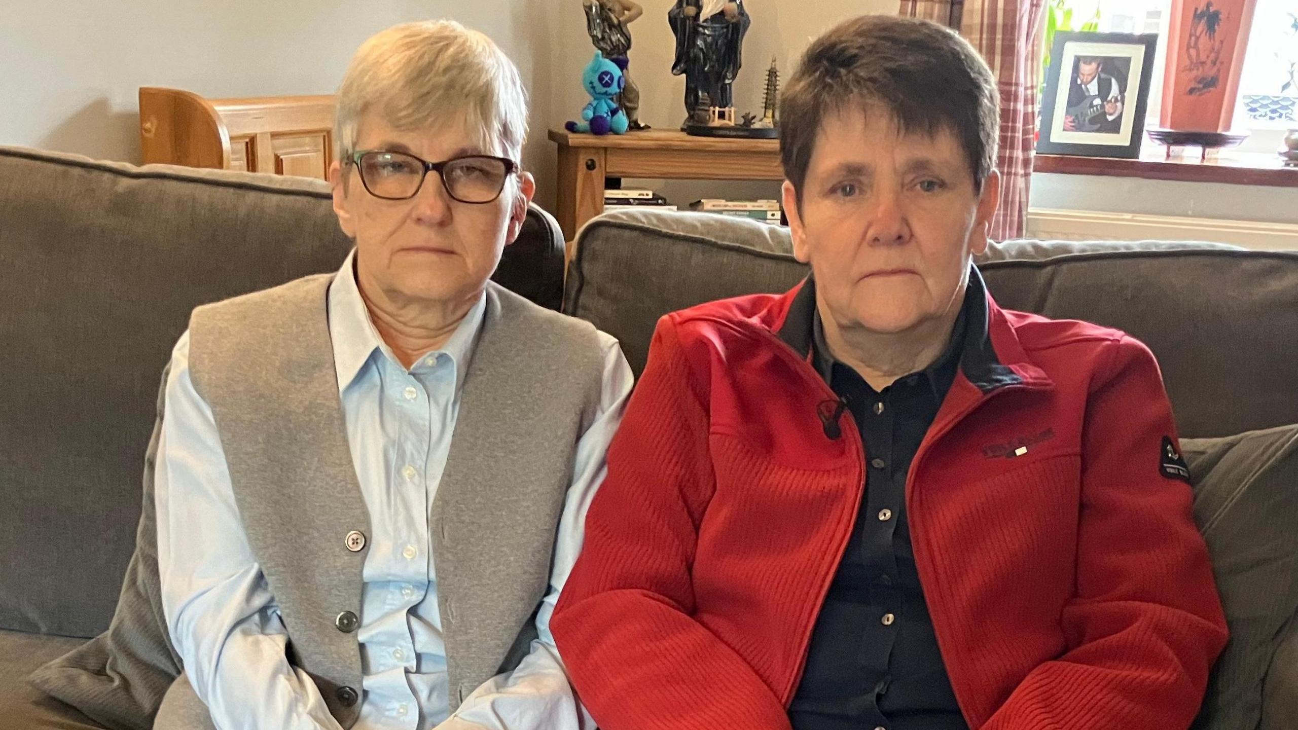 Two women sit next to each other on a brown sofa. The lady on the left, Sarah, is wearing a blue shirt, beige vest and glasses. She has short cropped hair. Karen who is sitting to  the right of her is wearing a red jacket and black shirt. She has brown short hair