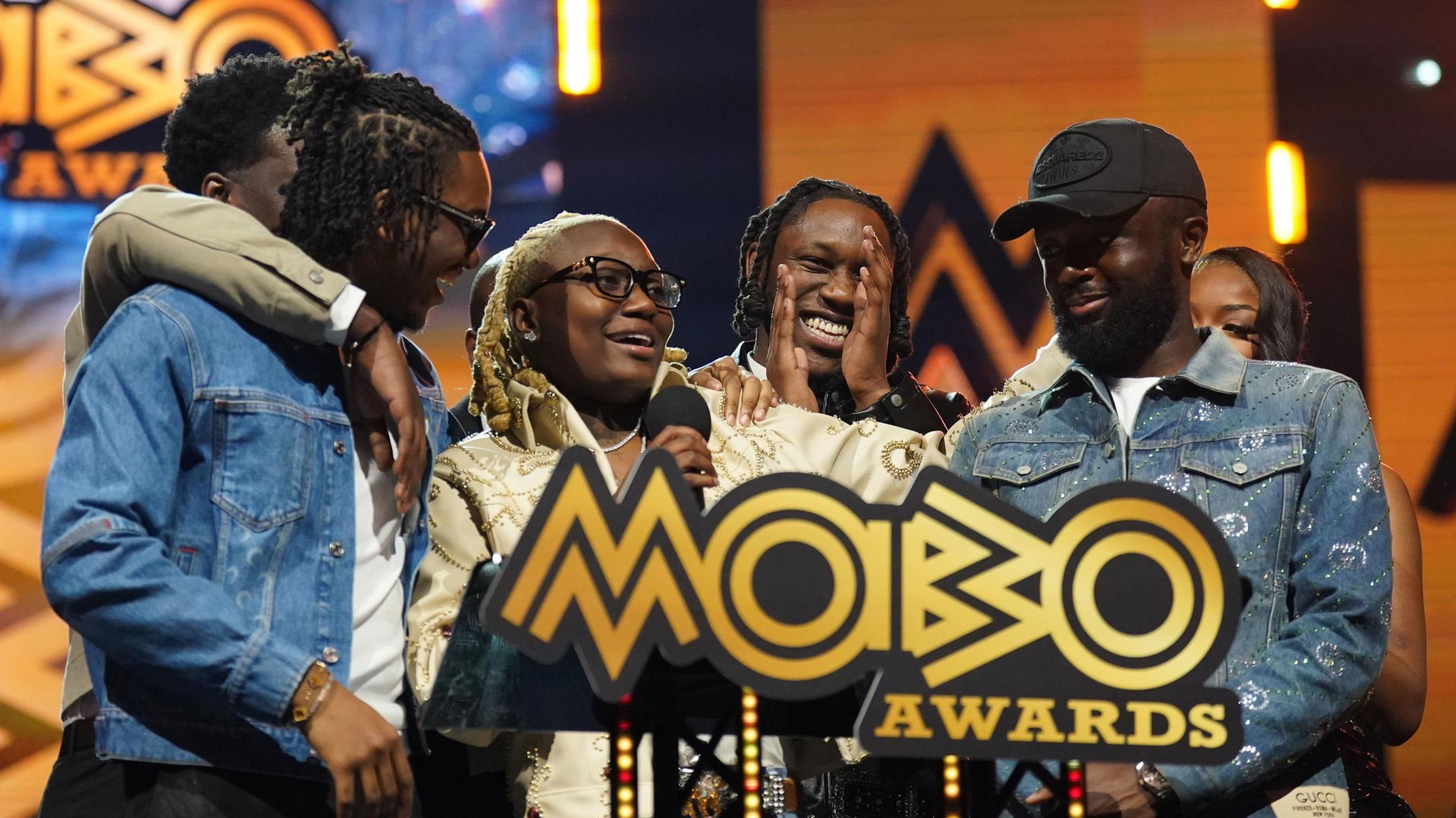 Darkoo (centre) accepts her award for best female act. She's surrounded by some supporters and looks emotional as she holds a mic.