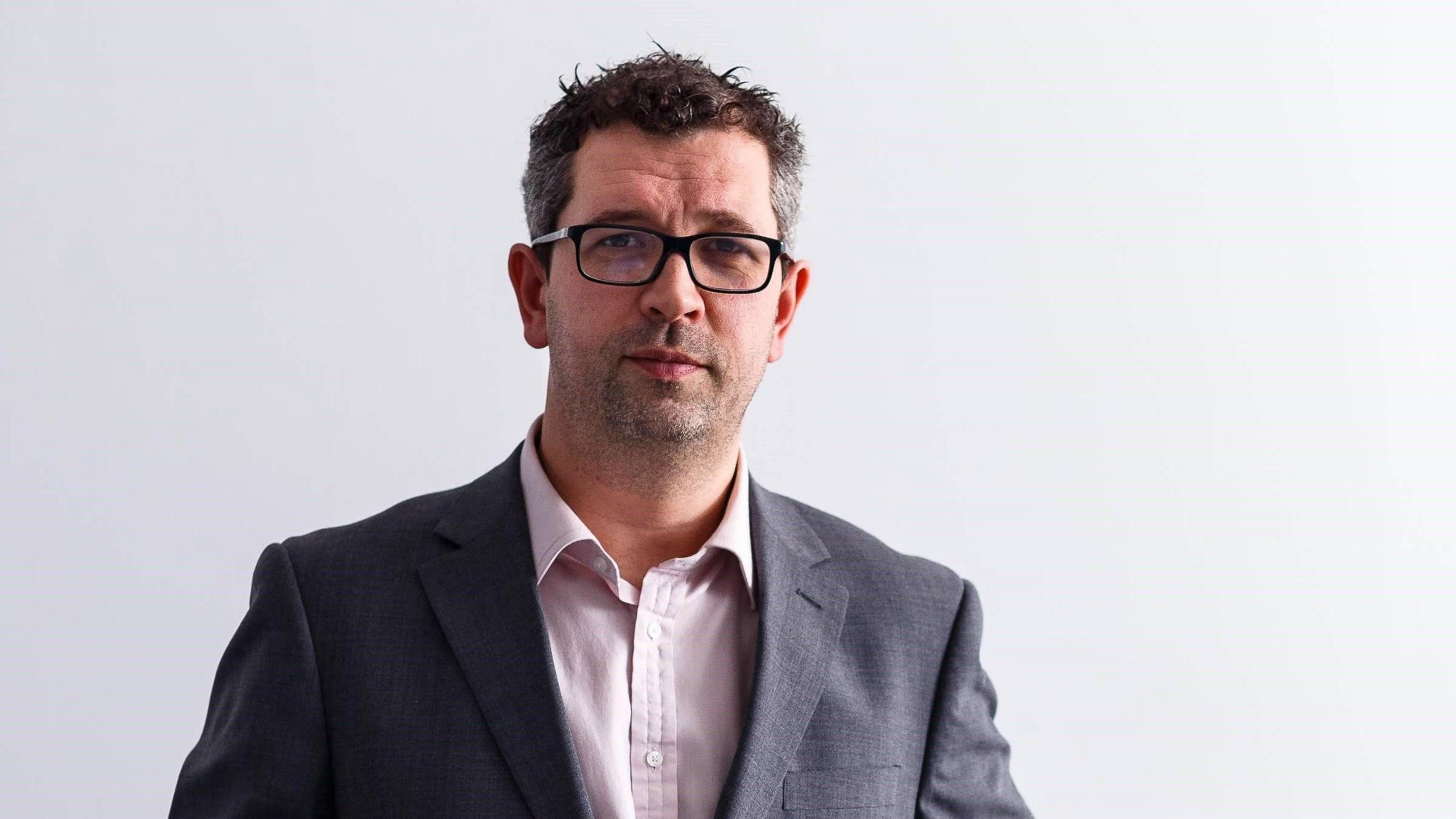 A professional headshot of Dan Panes. He stands against a white backdrop and wears a grey suit jacket and a white shirt. He has short dark brown hair and wears black rimmed glasses.