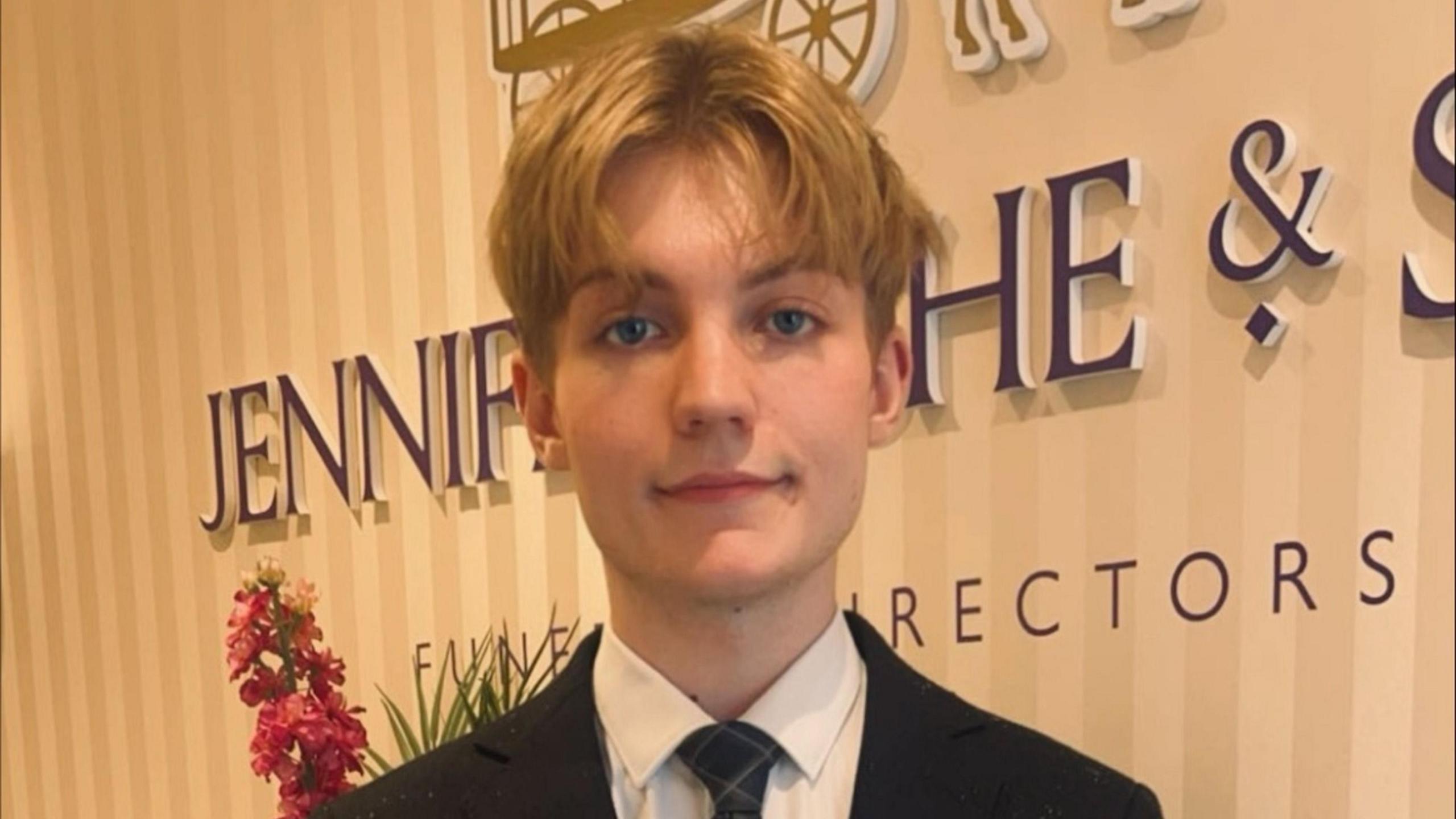 A young man with blonde hair smiles at the camera, in front of a funeral director's company sign