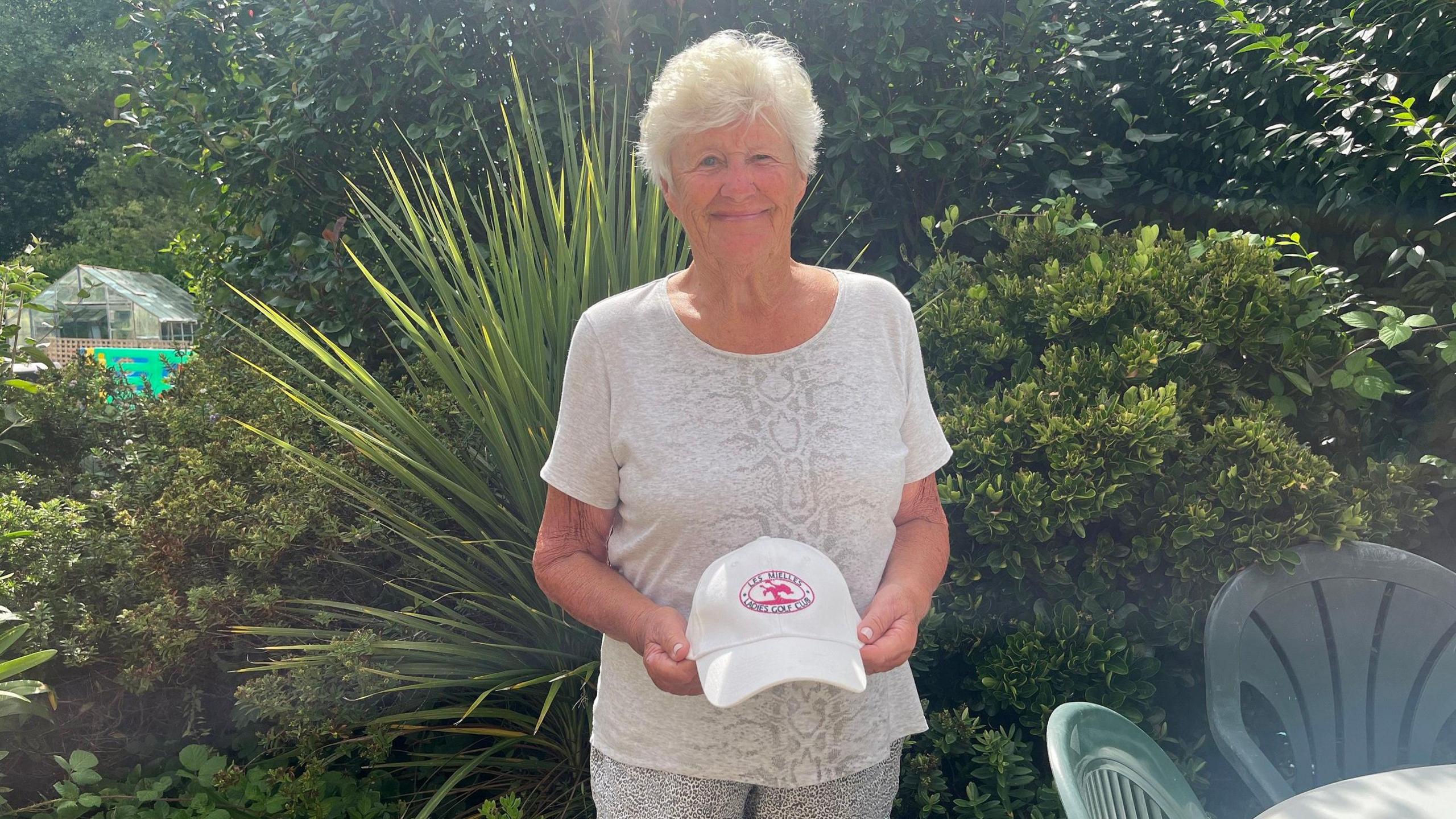 Margaret Vautier is smiling at the camera and she holds a golf club cap in her hands and is standing in a sunny garden with a table and chairs to her left and a greenhouse further down the garden