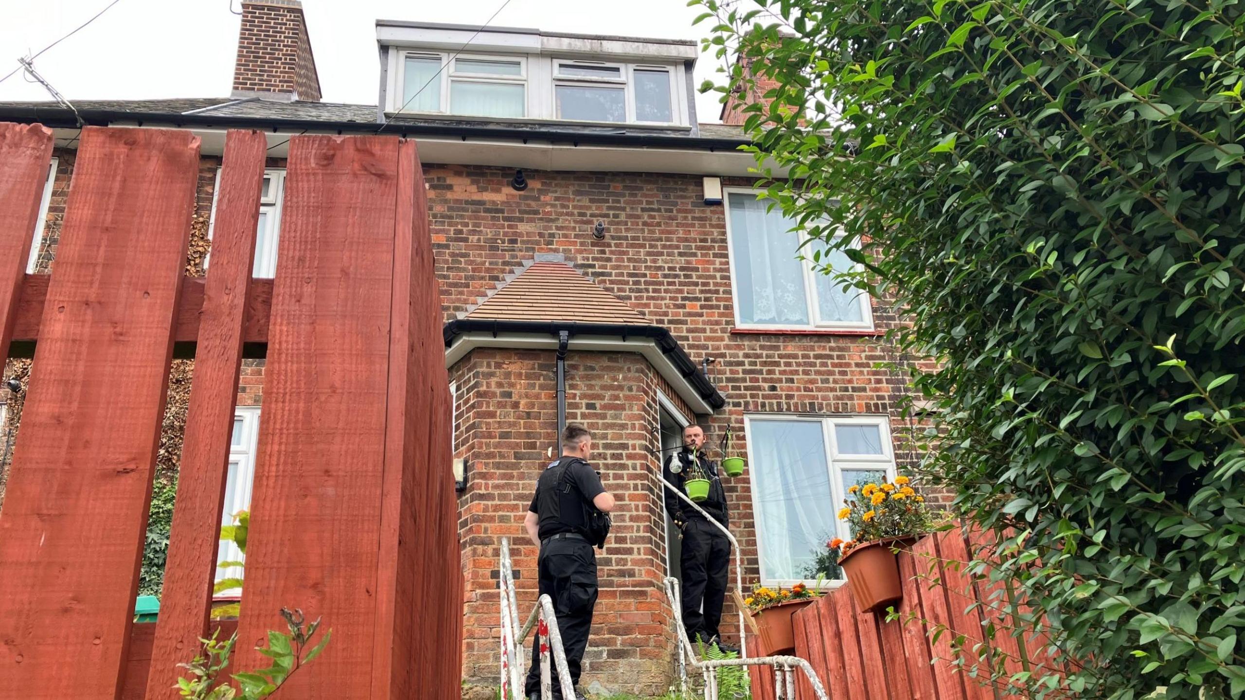 A police officer is stood at the front door of a three-storey terraced house, with another officer standing outside nearby. Oranges fences with potted plants on them can be seen in the front gardens of the houses