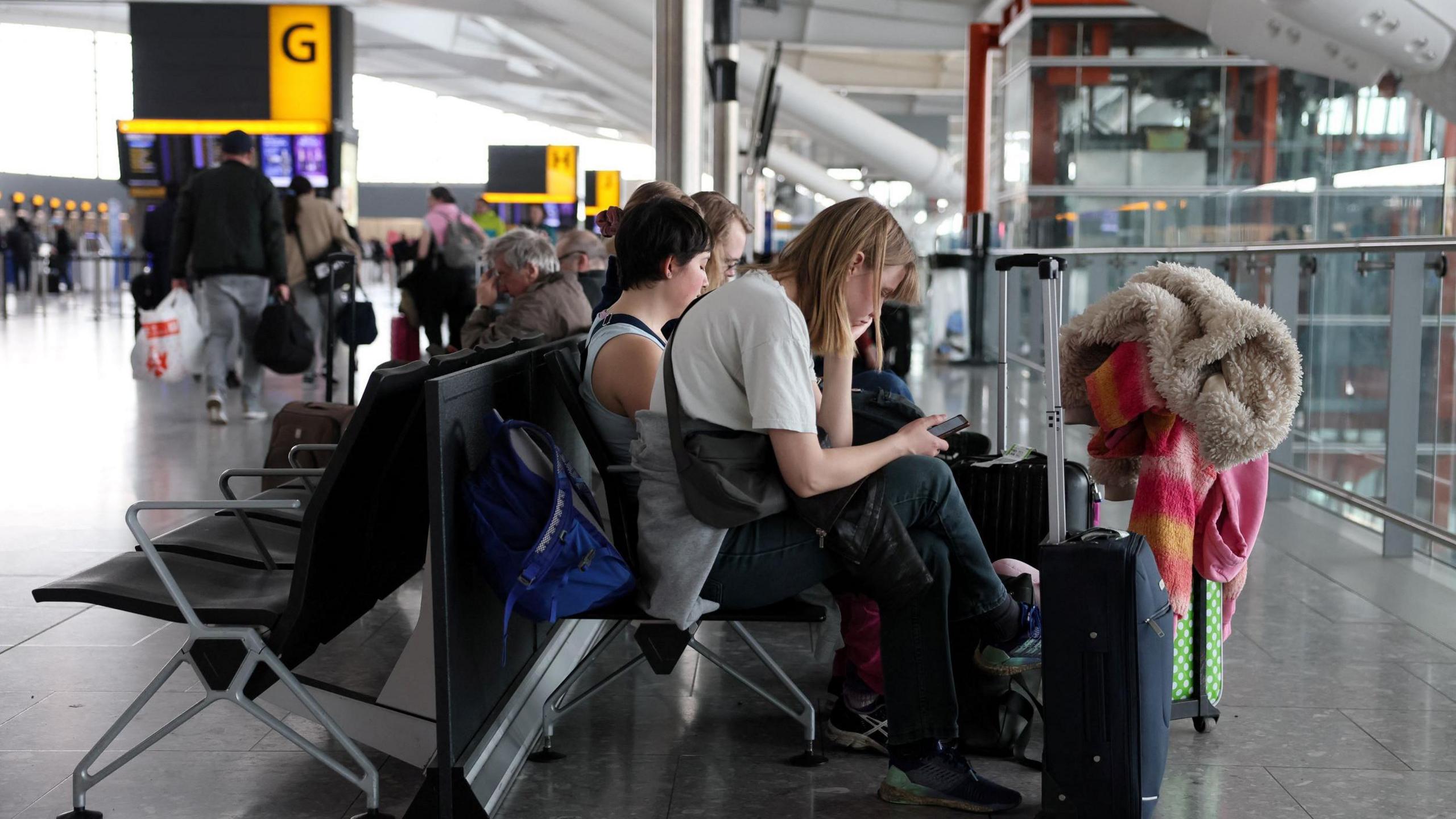 People sit on chairs at a terminal at Heathrow Airport. They are sat with suitcases in front of them.