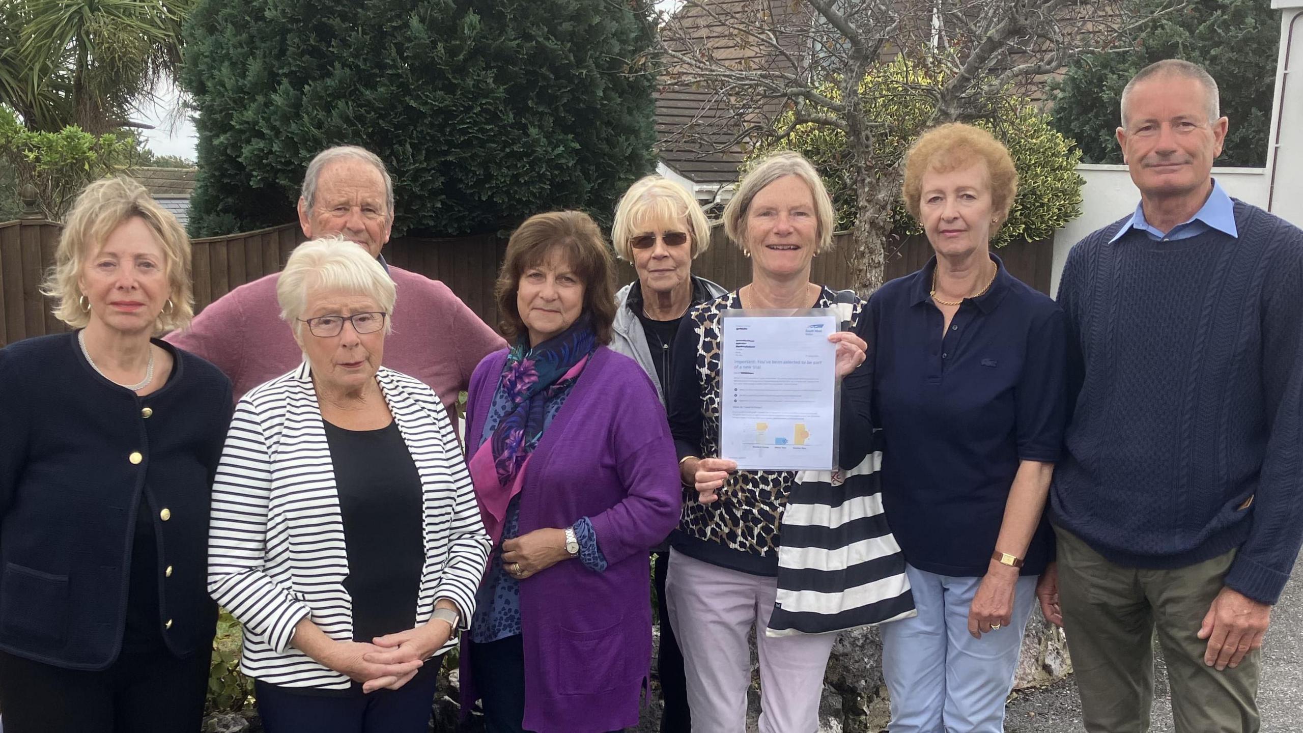 Eight people standing on a surfaced driveway in front of a low brick wall. They are all looking at the camera and one woman is holding a letter on an A4 page up but the contents of the page are too small to read