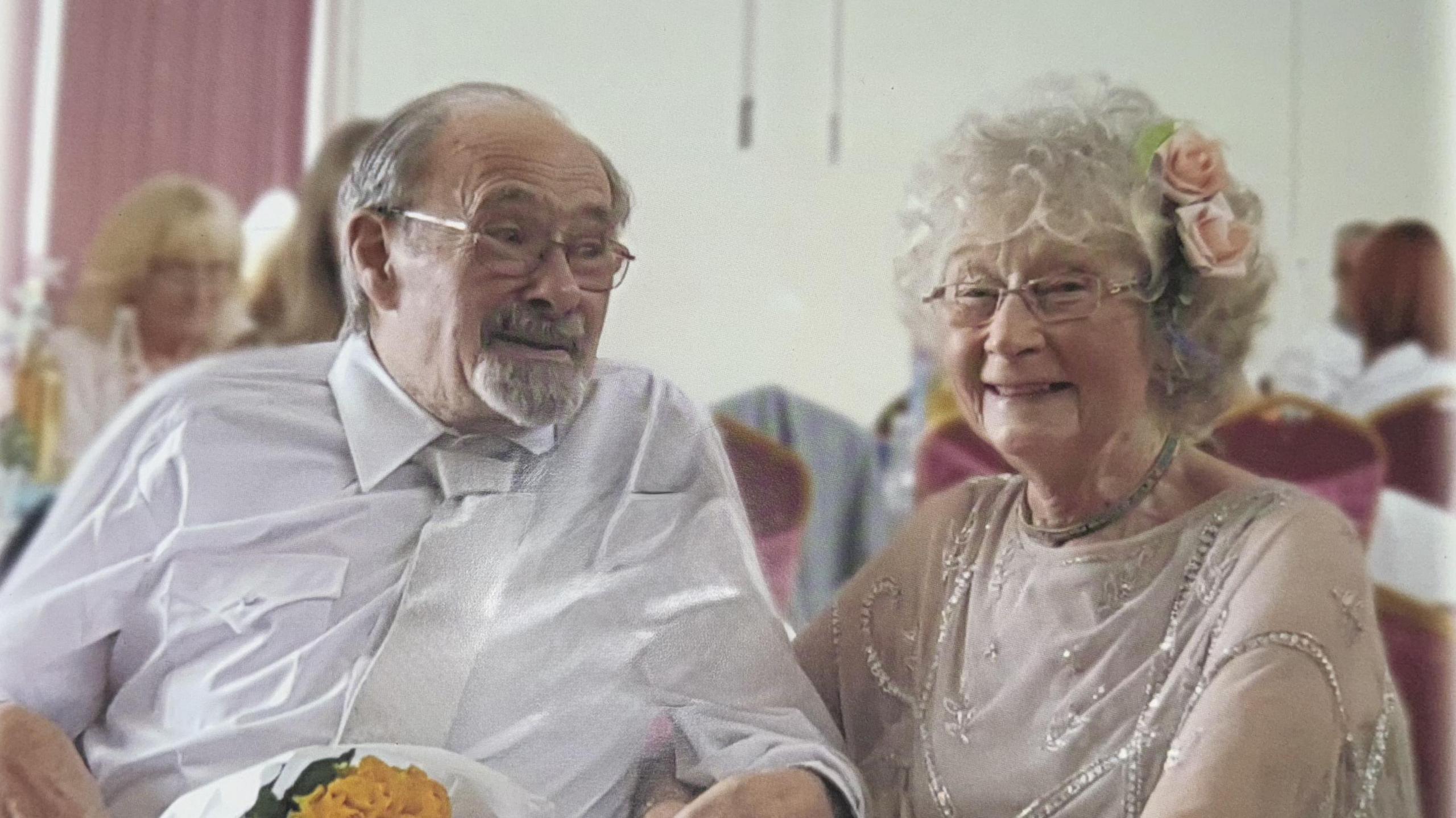 Man in shirt and tie in glasses on the right. He's holding some flowers in his hand. He's golding the hand of the women on the right. She has glasses and curly hair. She has a flower in hair and is smiling.