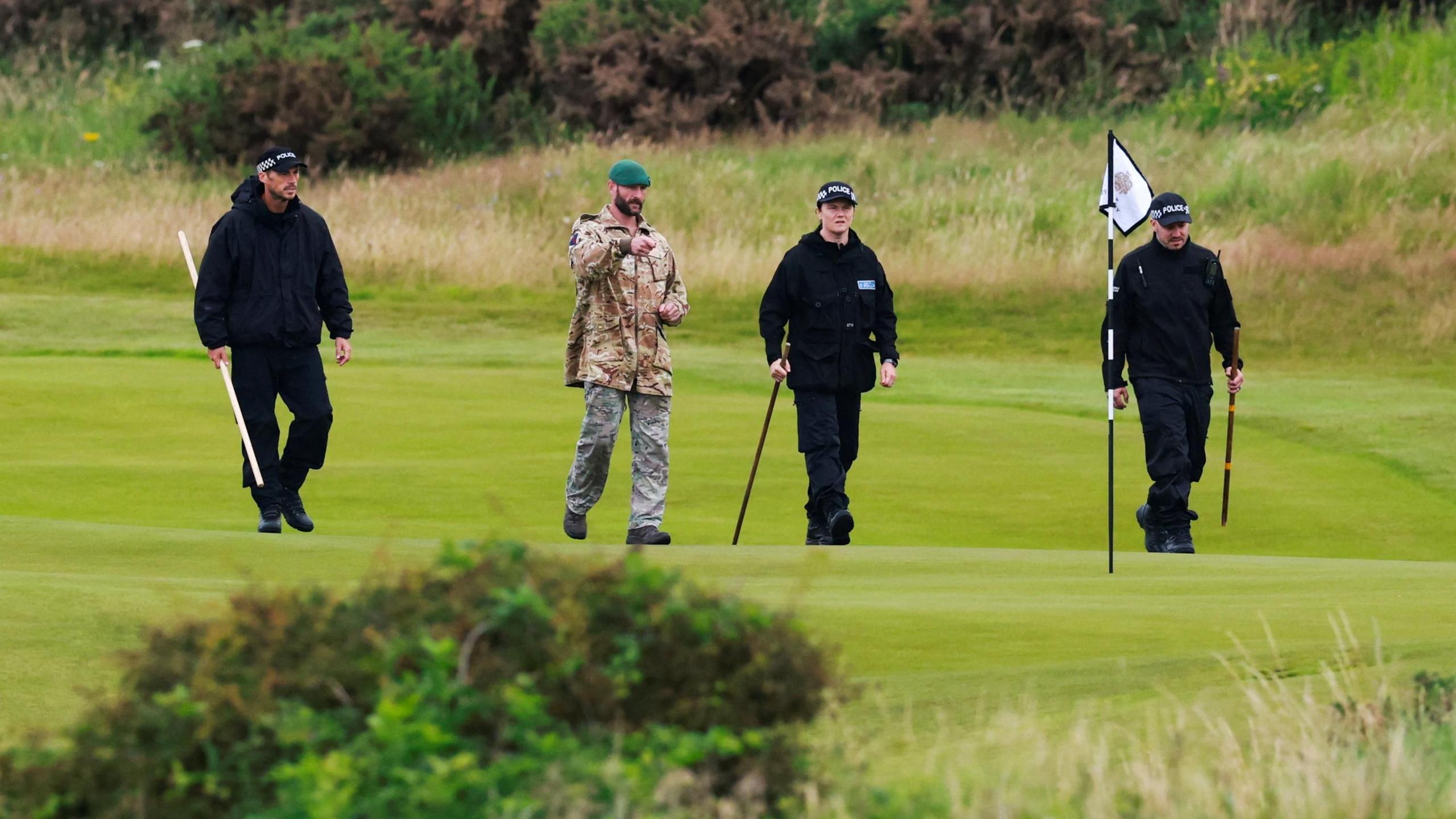 Three police officers carrying poles and one member of military personnel walking on the golf course next to one of the greens. 