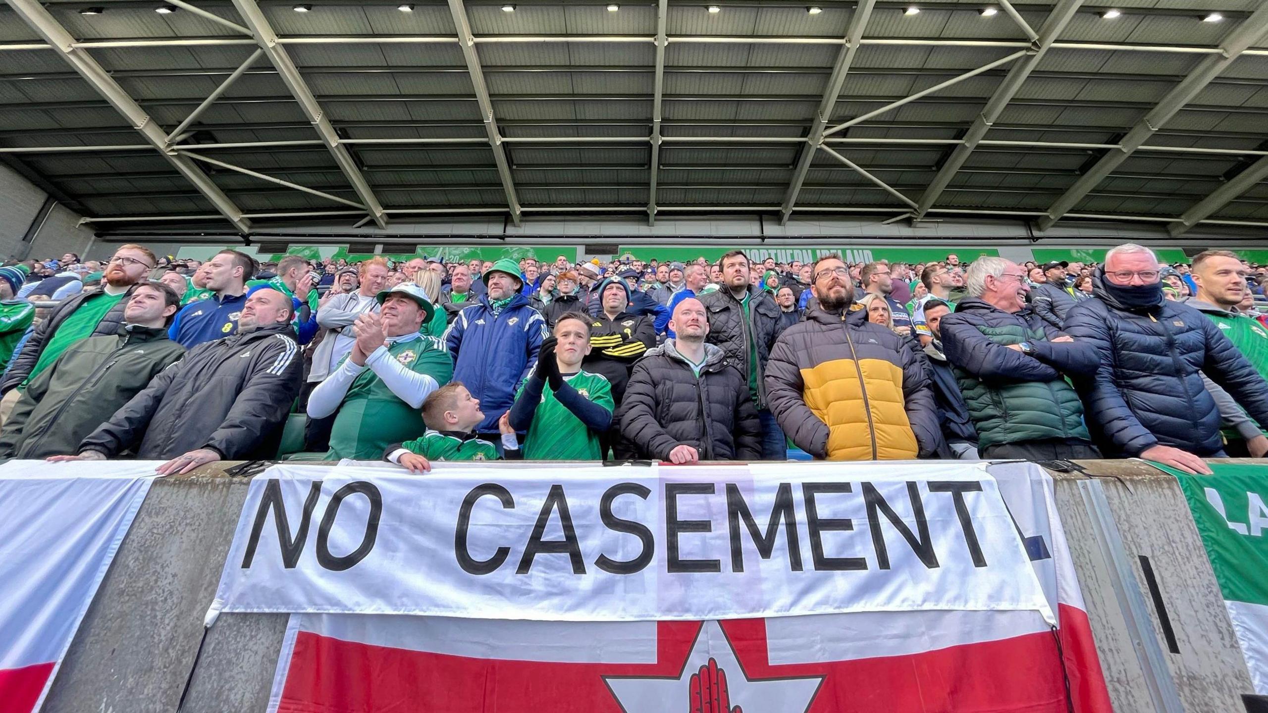 Supporters in the stands, many wearing Northern Ireland football jersey's. There is a wall in front of the front row of seats. On it is an Ulster Banner flag, and a sign saying 'no casement'. 