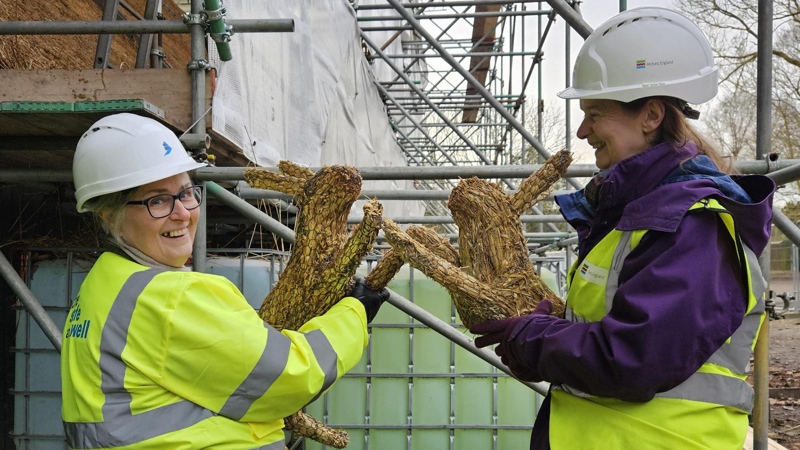 Two women wearing high-viz and hard hats hold thatched hares.