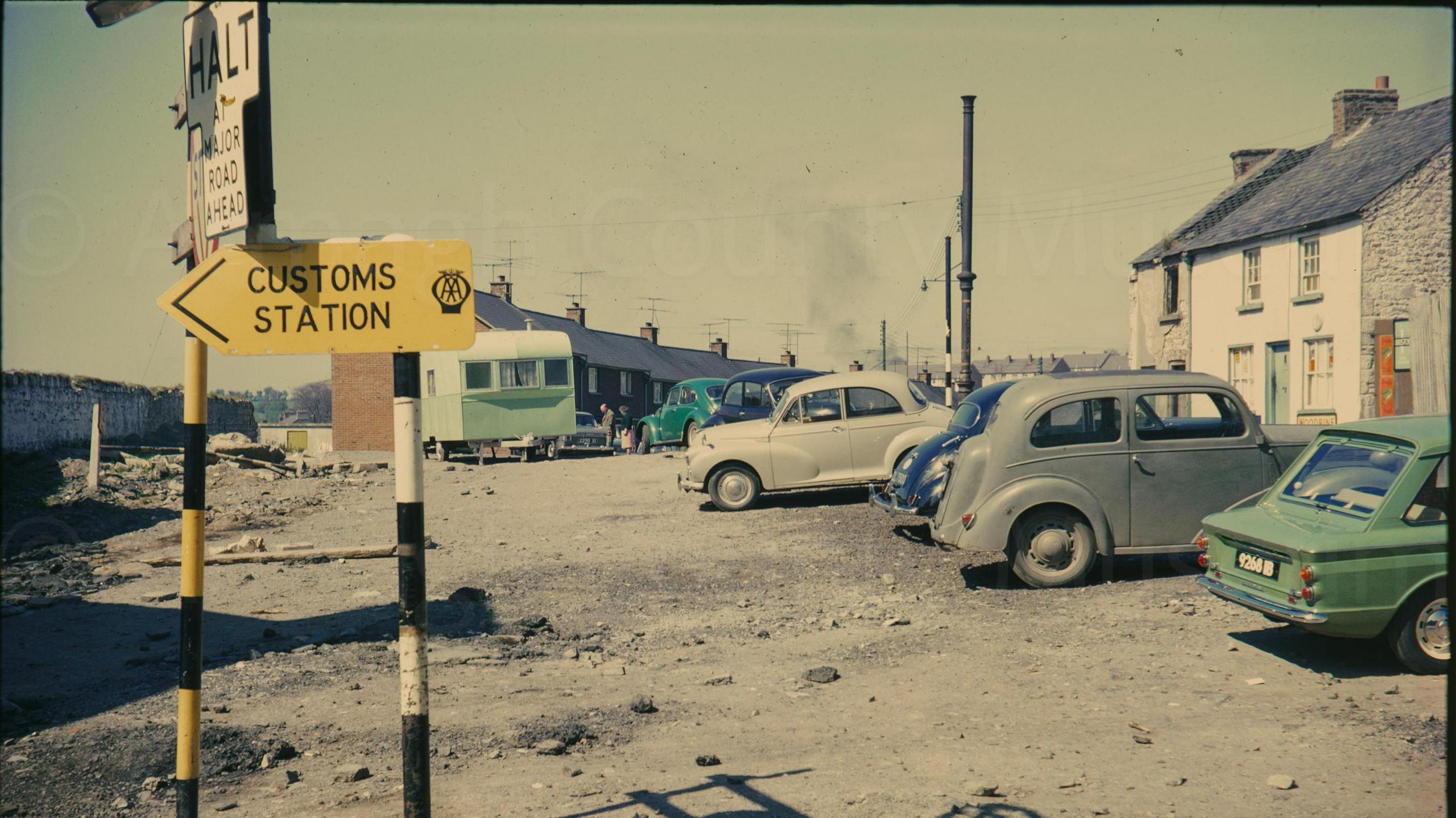 A digitalised photo from a film of a car park, Green, grey and white cars are parked. Looking west from Irish Street corner several cars are parked on the cleared ground near the camera and the terrace of new houses a partially visible in the background. An old and partially derelict house is on the right of the frame and a yellow finger post sign reads "Customs Station" May 1967.