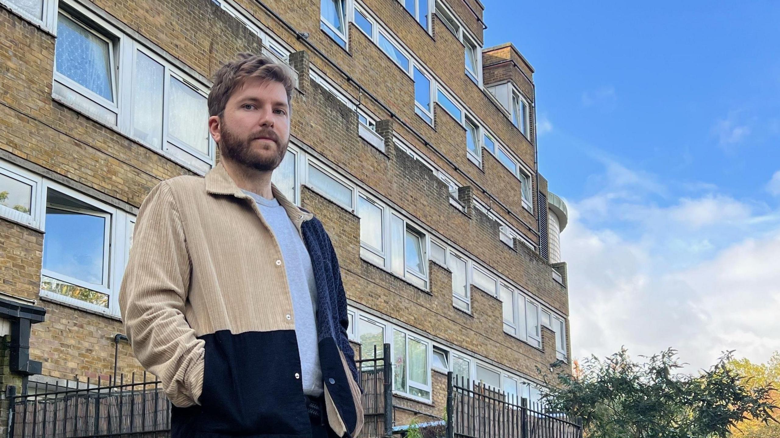 An image of Max Templer, a young man with short brown hair and a beard wearing a patchwork jacket and grey T-shirt. He is stood in front of his brick block of flats