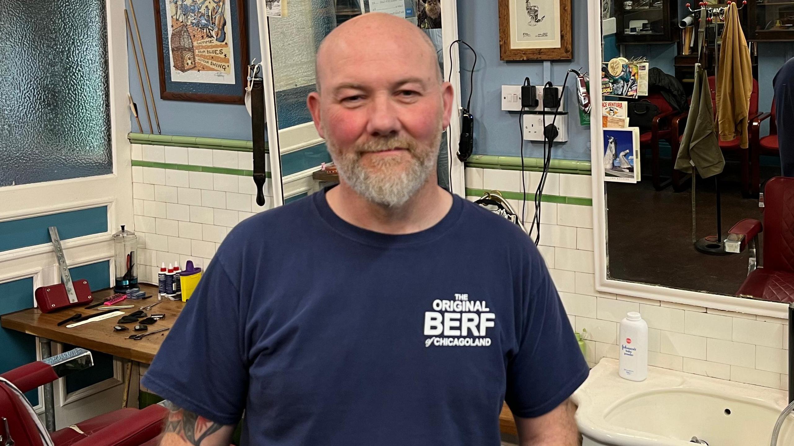 Barry Whincup. He is wearing a blue T-shirt and is bald with a greying beard. He is standing in front of his barber's chair at the shop in Newcastle. The walls are painted blue and have tiles lower down. There are old fashioned drawings in frames on the wall between the mirrors.