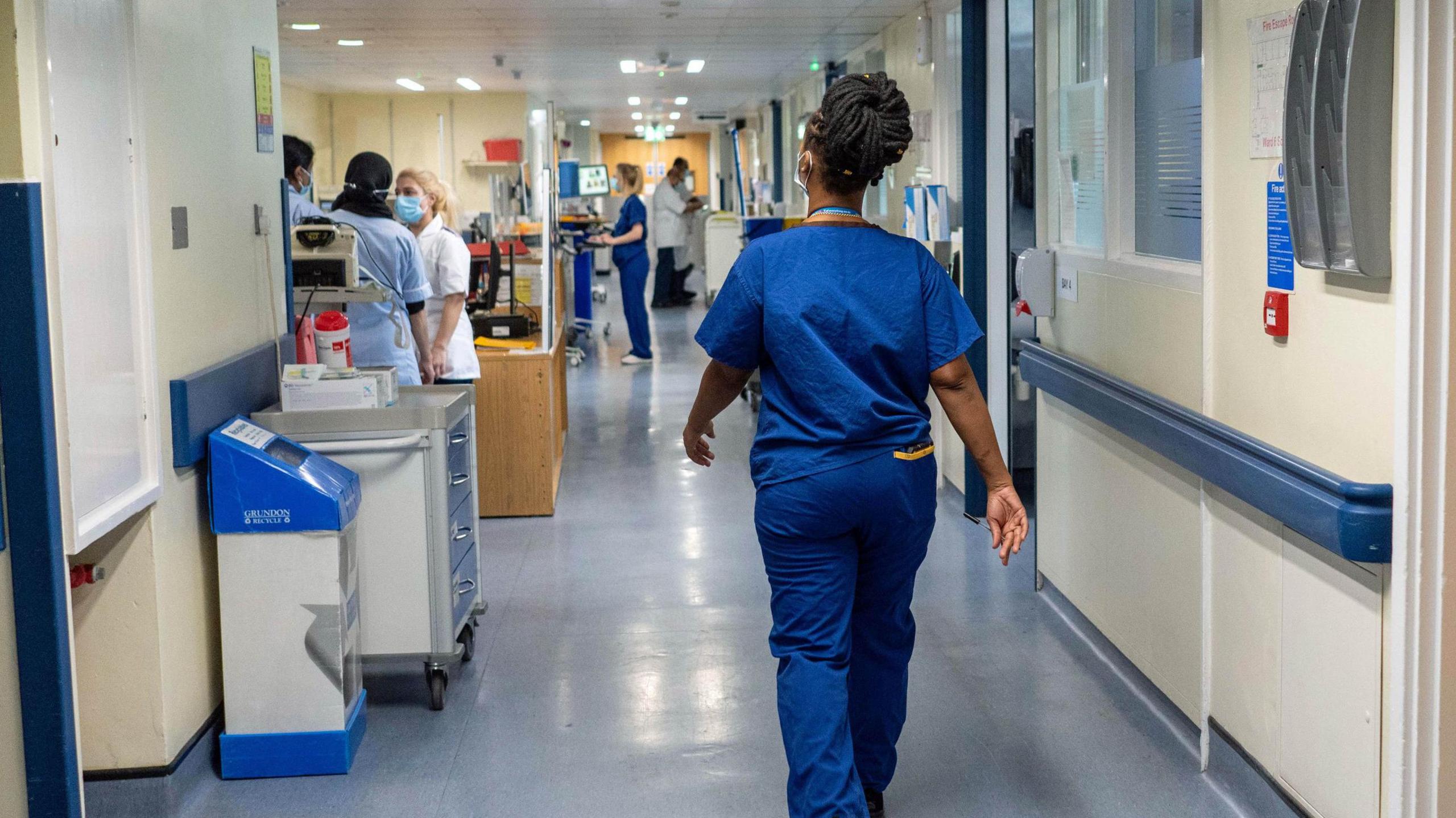 A doctor walks down a hospital corridor, as other medical staff gather further down.