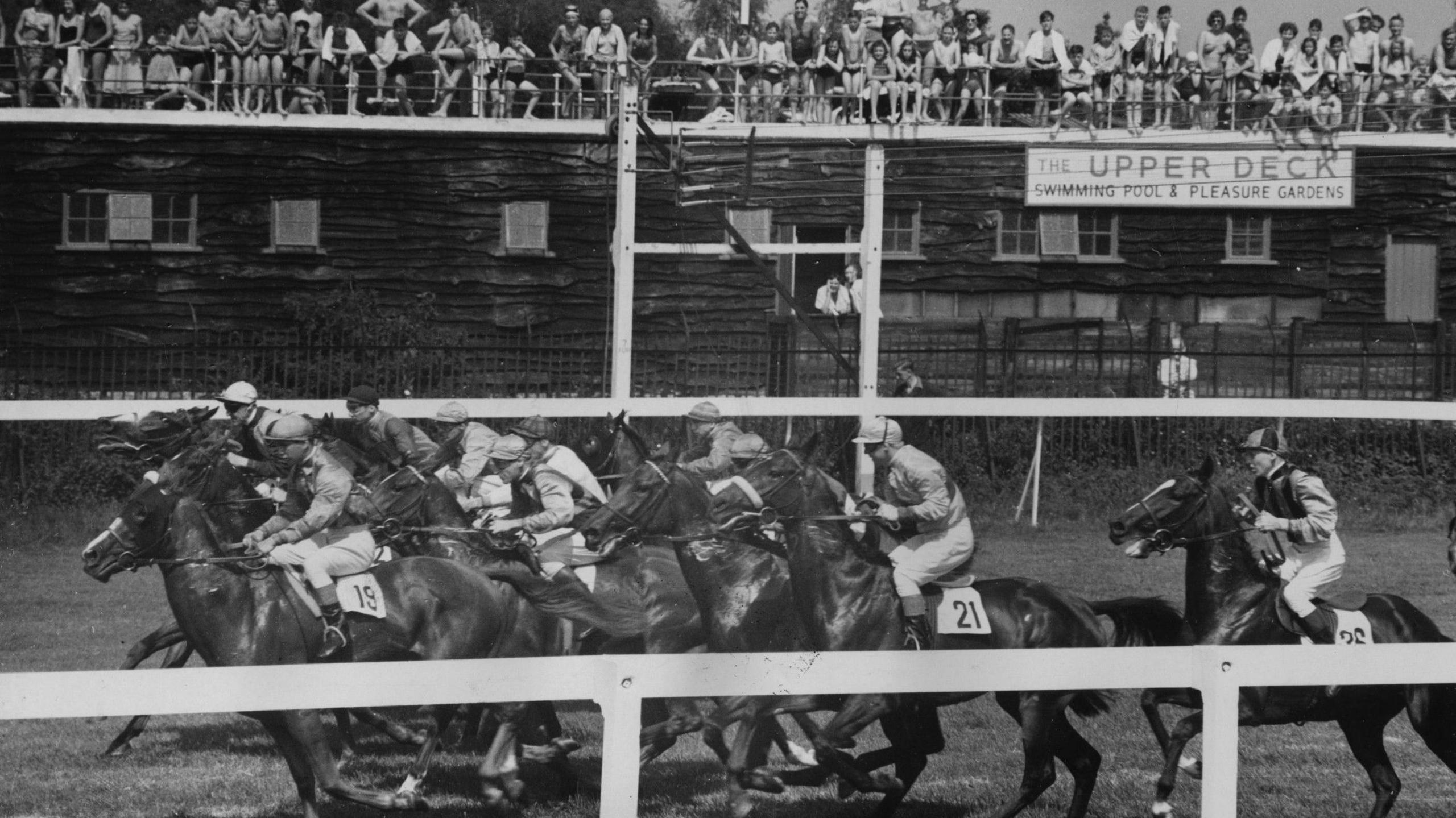 27th July 1956: Crowds on the upper deck swimming pool at East Molesey in Surrey watching the racing at Hurst Park racecourse. A large group of horses race close to the white rails around the inside of the track.