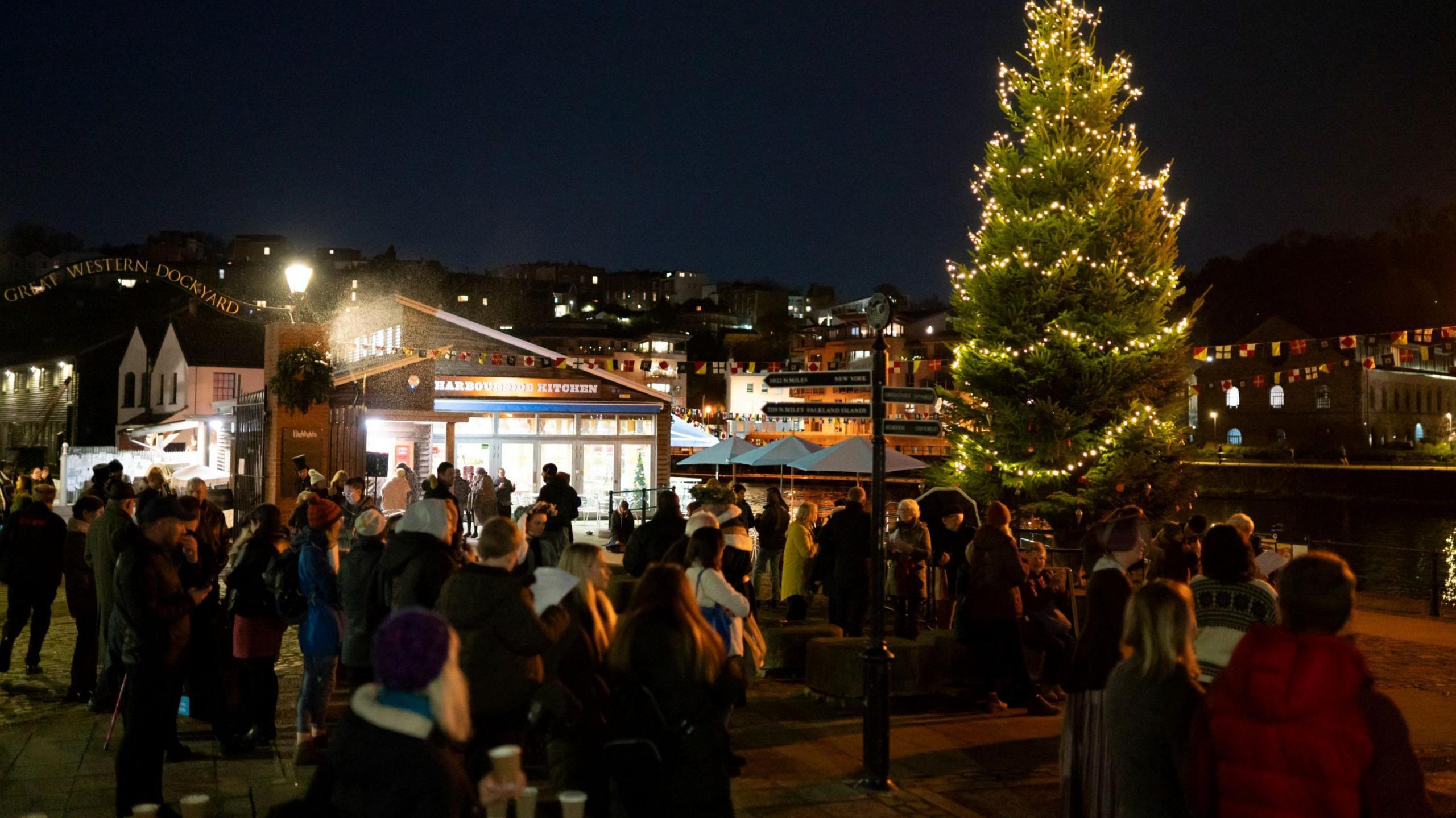 Dozens of people gather round a Christmas tree on the side of Bristol Harbour next to the SS Great Britain. The picture is taken at night and the tree is illuminated with bright lights. Clifton housing can be seen in the background