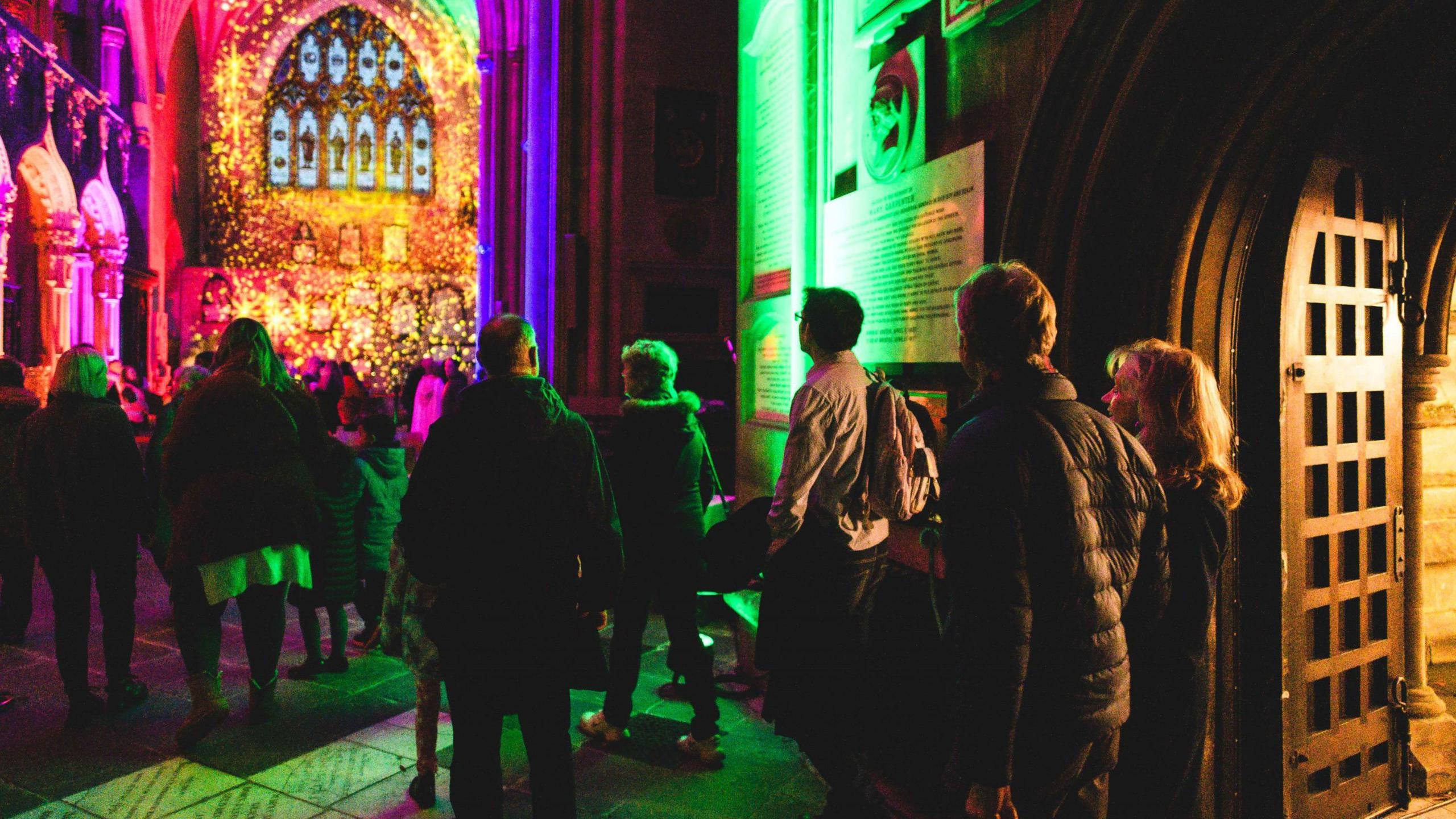 People walk into Bristol Cathedral at night, heading towards the light display called Luxmuralis which is lighting up the nave of the cathedral in the background with a mixture of yellow and purple lights