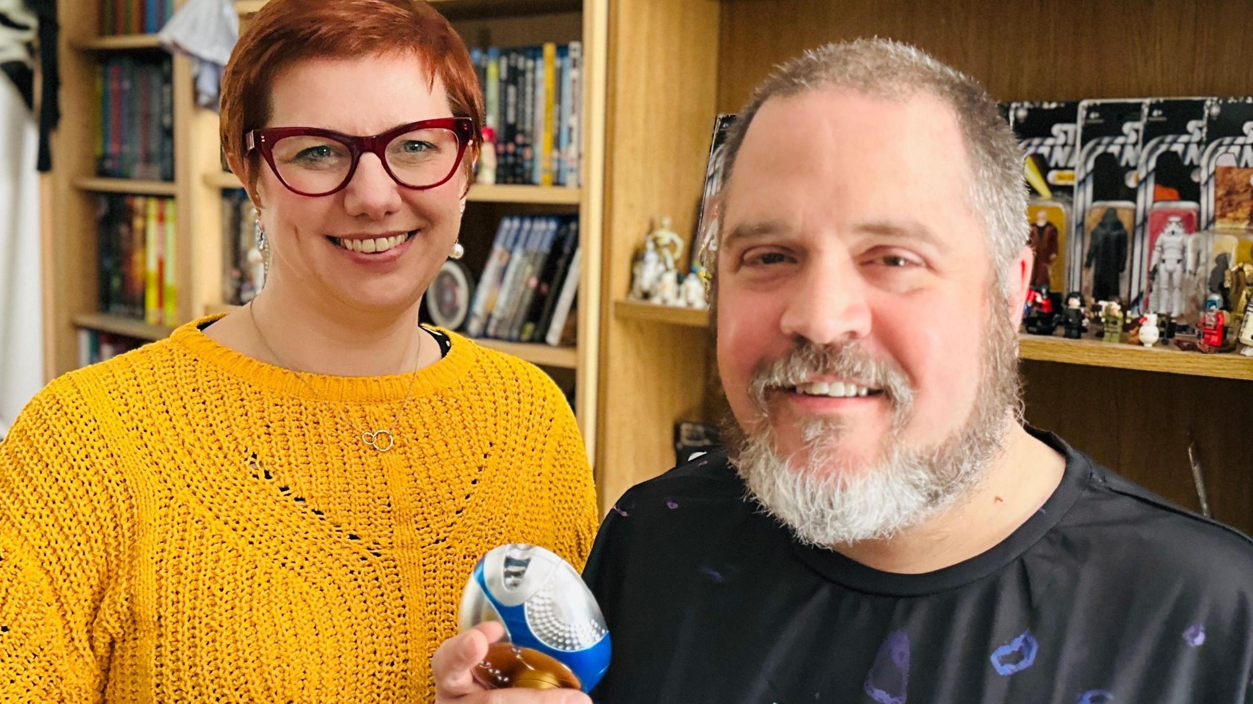 Emily Garside with short red hair and glasses, wearing a mustard yellow knit jumper, stood next to a man with short grey hair and a beard. He is holding a small piece of Doctor Who merchandise and they are stood in front of bookcases full of other fandom merchandise.
