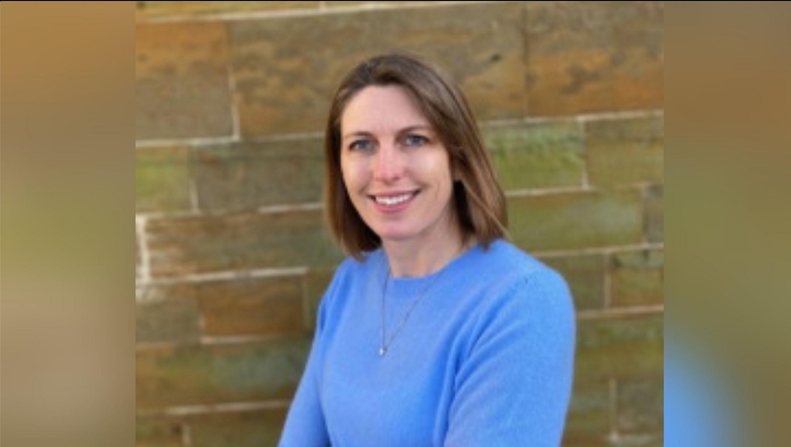 Louisa Fowler with brown hair wearing a blue top in front of a wall