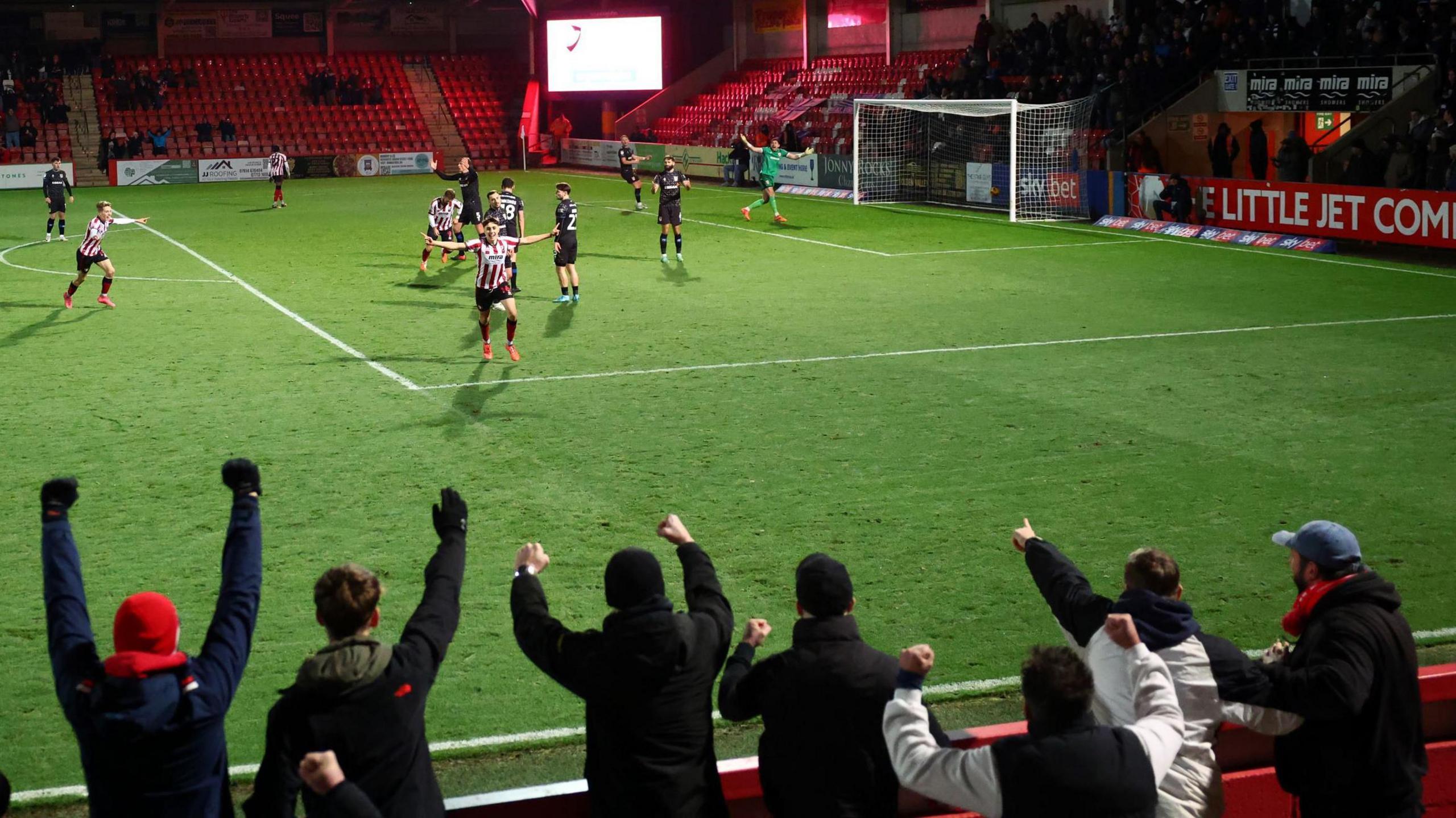 Cheltenham Town players run towards their fans to celebrate after Joel Colwill scored the only goal of the game against Tranmere at Whaddon Road. The match is being played at night so the floodlights are on and fans in the foreground have their arms in the air in celebration