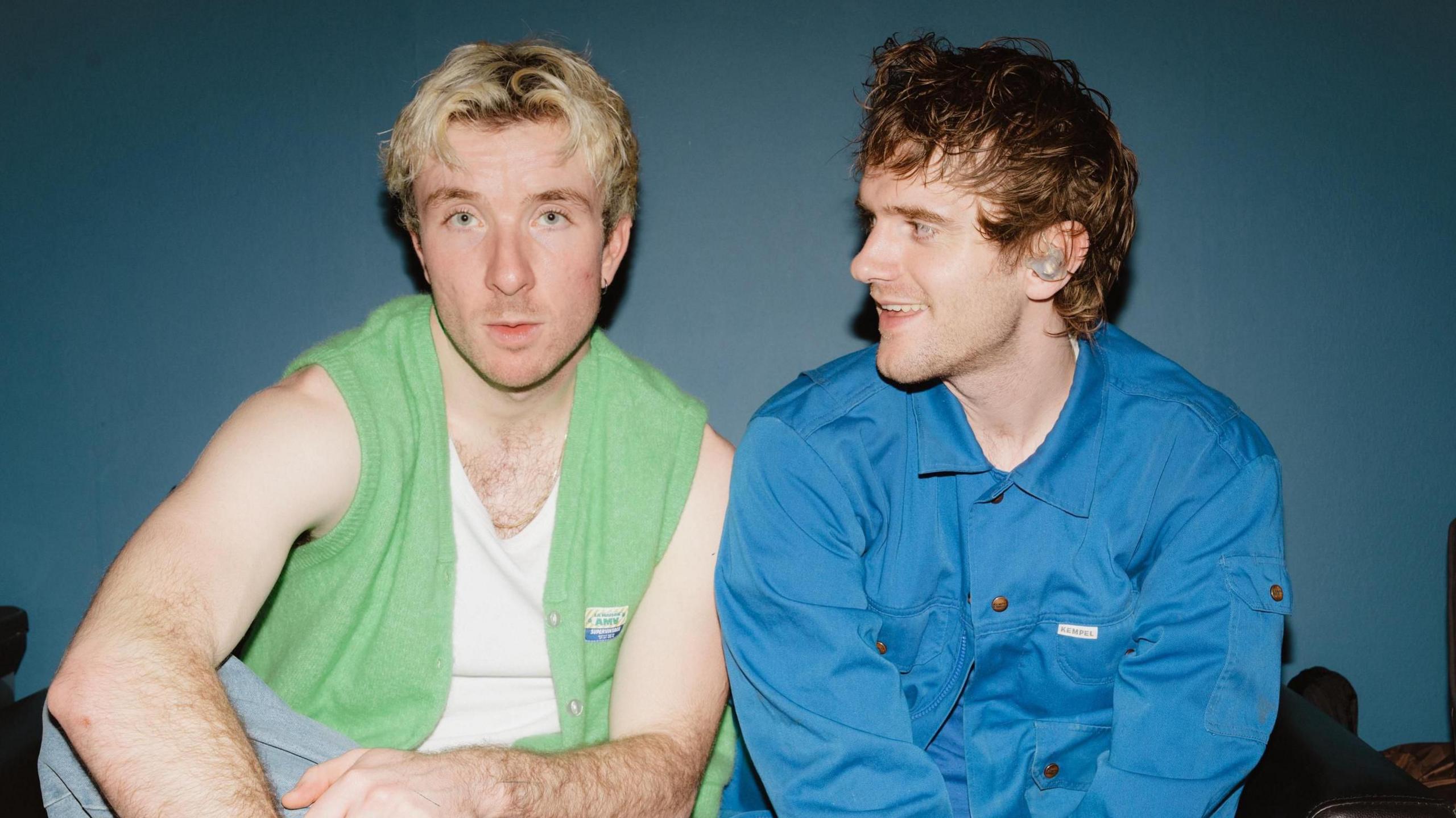 Good Neighbours made up of of (L-R) Oli Fox and Scott Verrill. Oli wears a green vest-cardigan over a white vest top, his hands in his lap as he sits cross-legged. Scott sits beside his bandmate and looks across at him, smiling, while wearing a blue overshirt. 