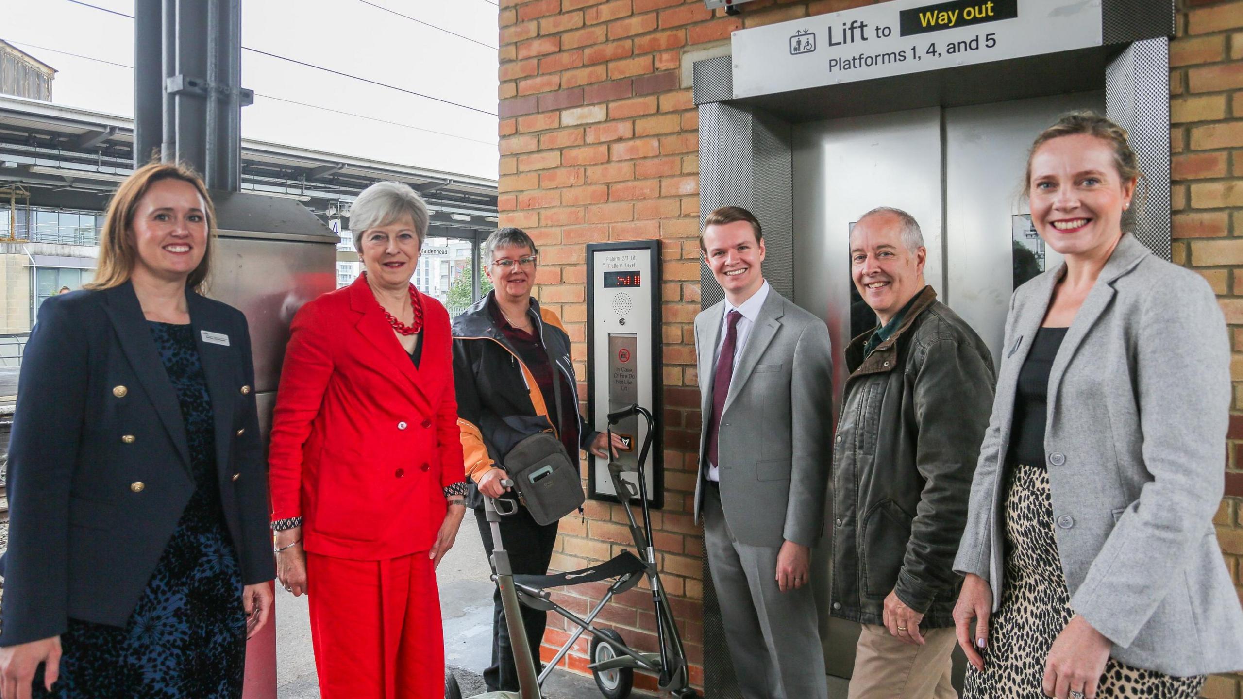 A group of six people stand in front of the newly opened lift, including Lady Theresa May who wears a bright red two-piece suit, MP Joshua Reynolds and Simon Werner, leader of Windsor and Maidenhead Borough Council