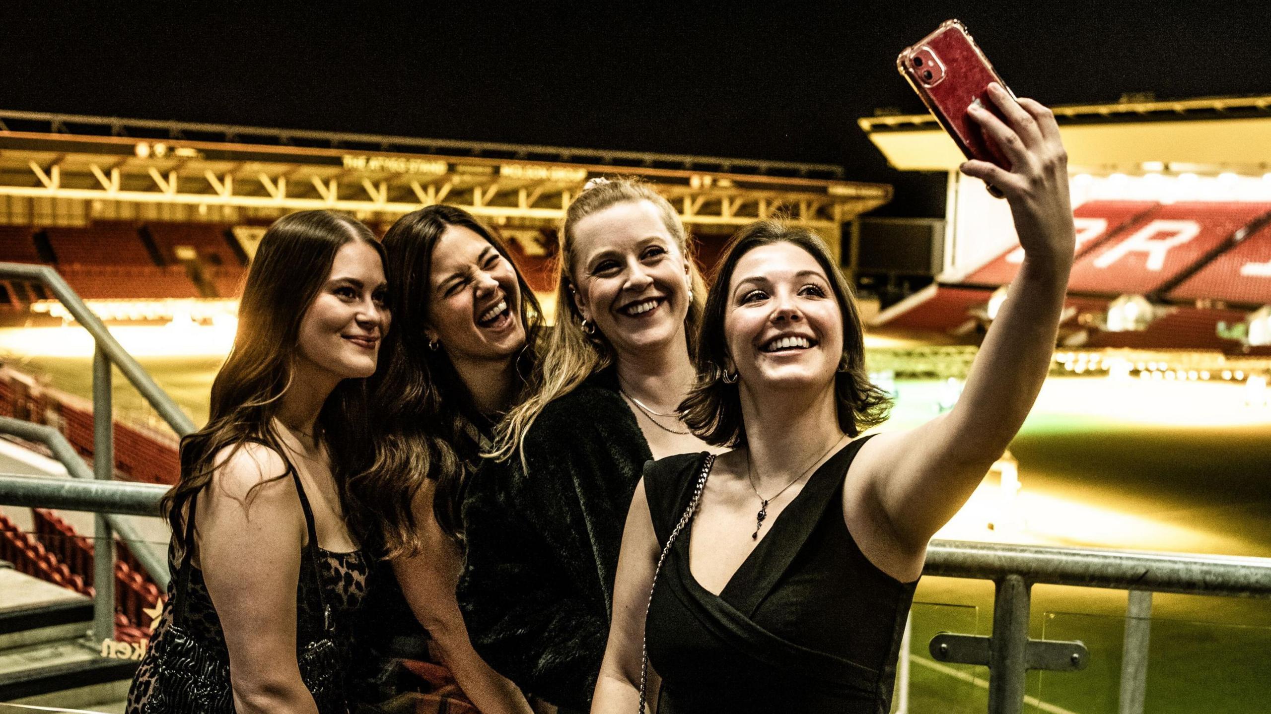 A group of four women in smart dresses pose for a selfie at the South West Tech Awards at Ashton Gate. They are outside on a balcony with the stadium and pitch in the background at night. They are all smiling and posing for the phone camera