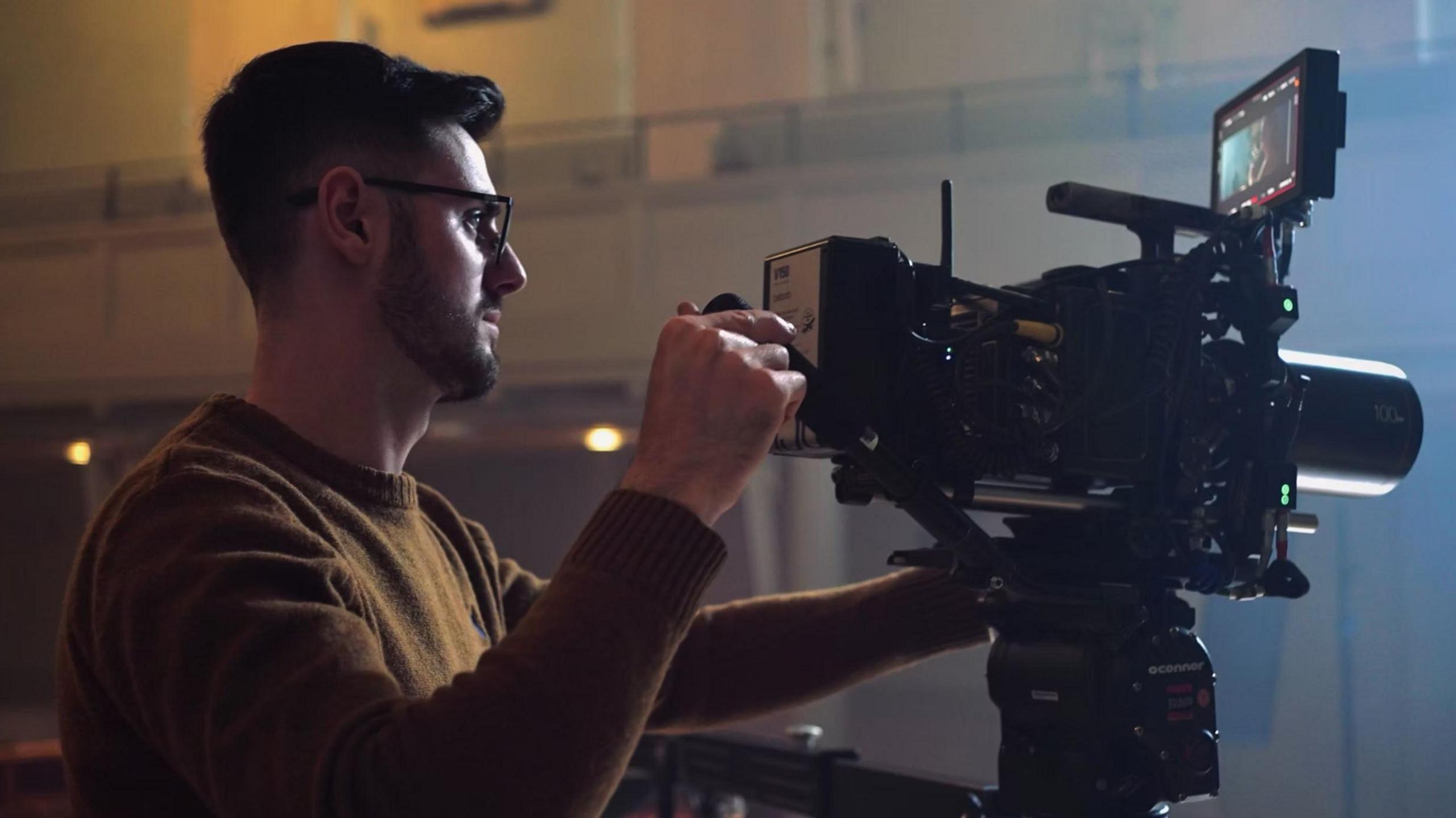 A man is wearing a brown jumper and black glasses. He is holding a large camera and filming a room.