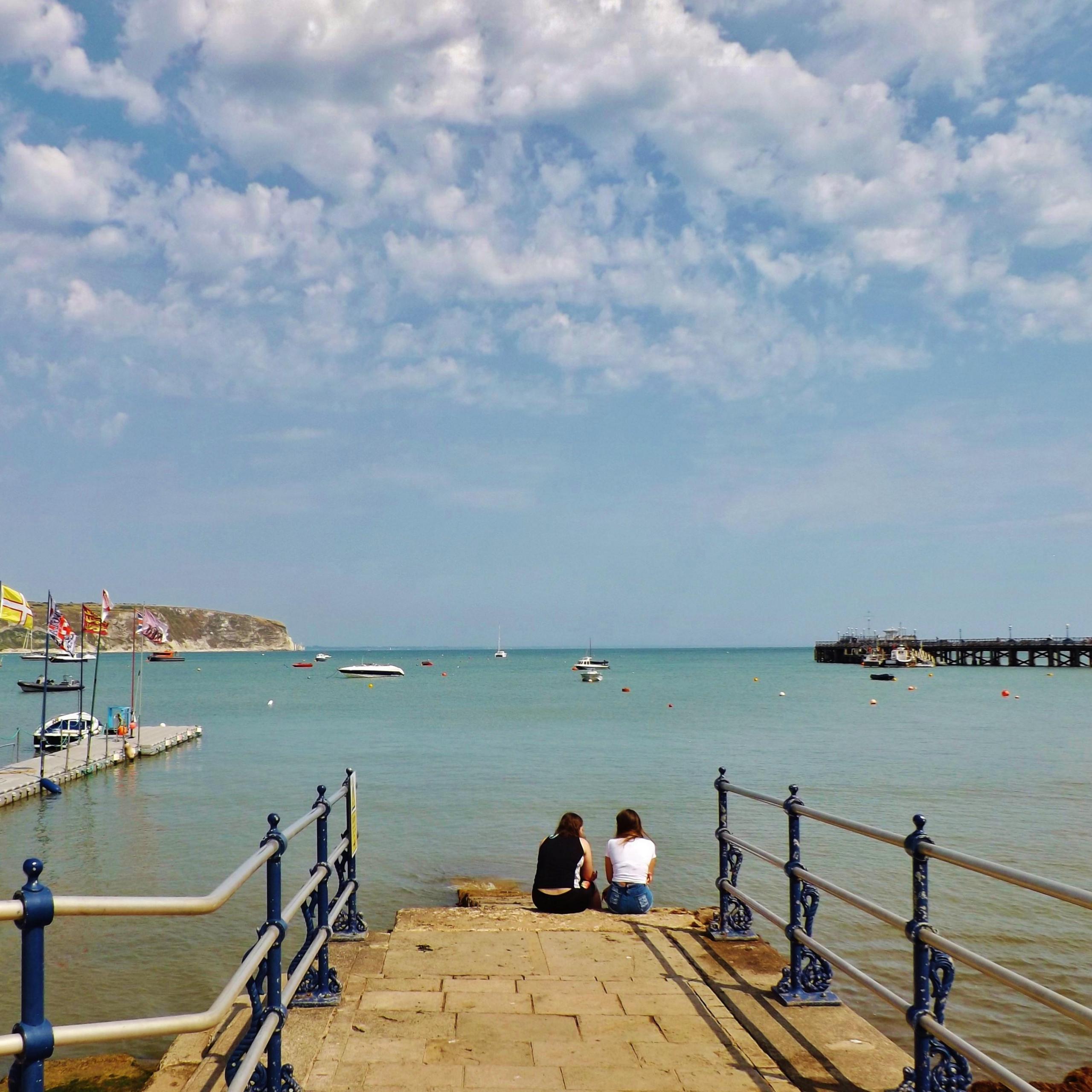 Two people sitting on the end of a short slipway with a pontoon, harbour and pier in the background