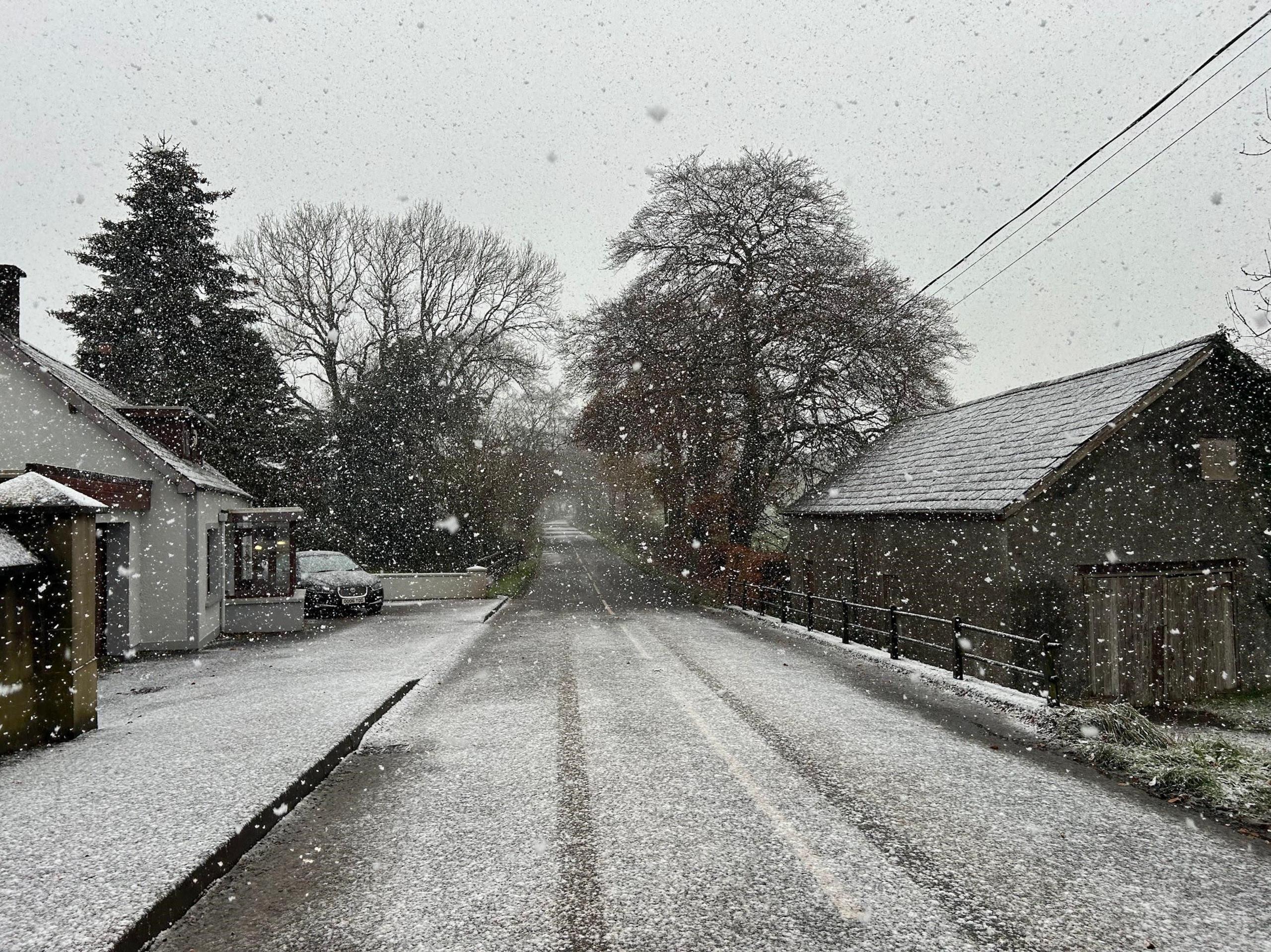 SA snow covered street in Ballymena. Snow is falling and tyre marks can be seen on the road. A parked car, outside a house on the left, has a covering of snow