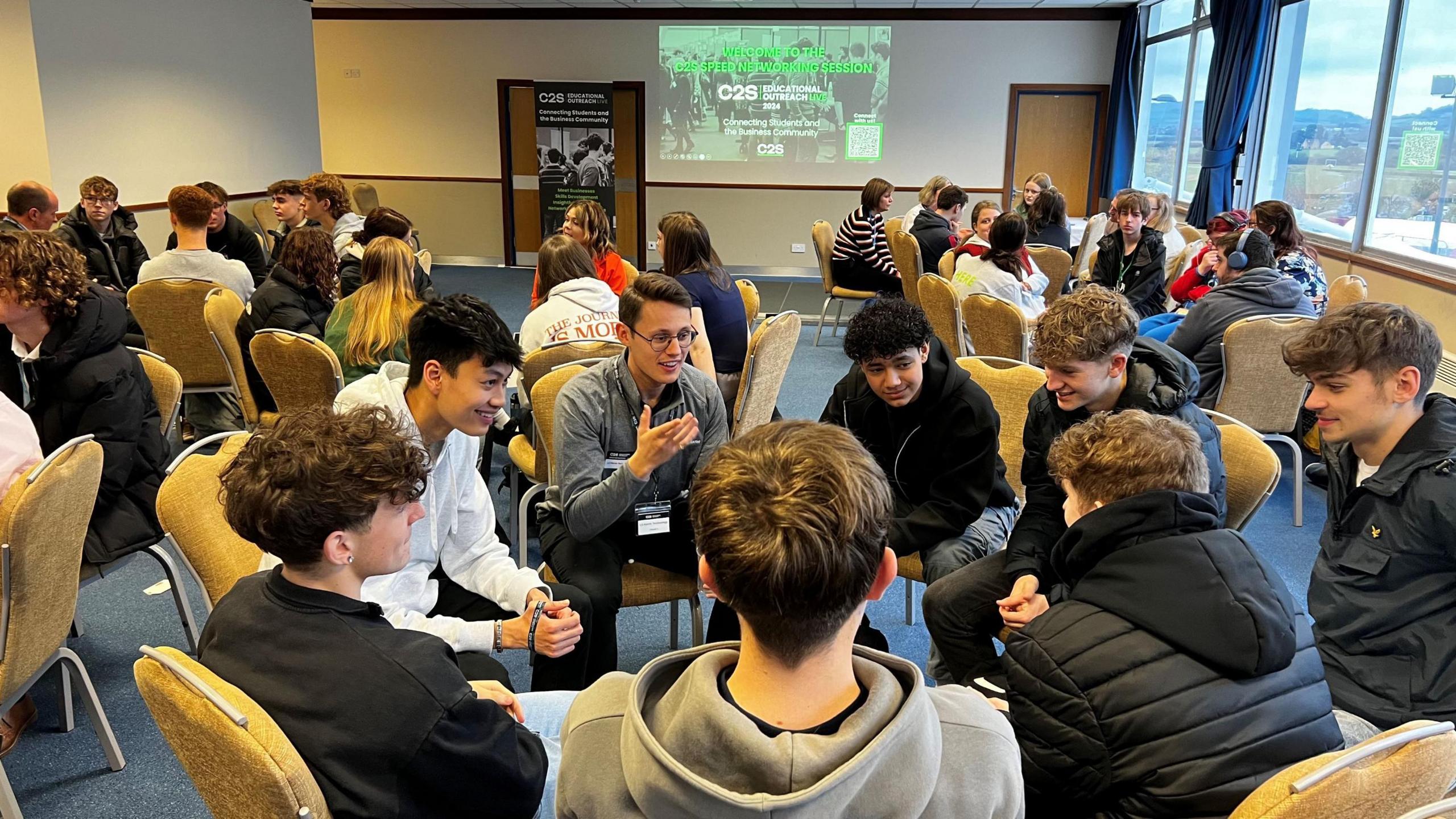 Dozens of students attending a careers fair at Cheltenham Racecourse sit in groups with their chairs facing each other, chatting. They are in a large room with plain beige-coloured walls and blue carpet