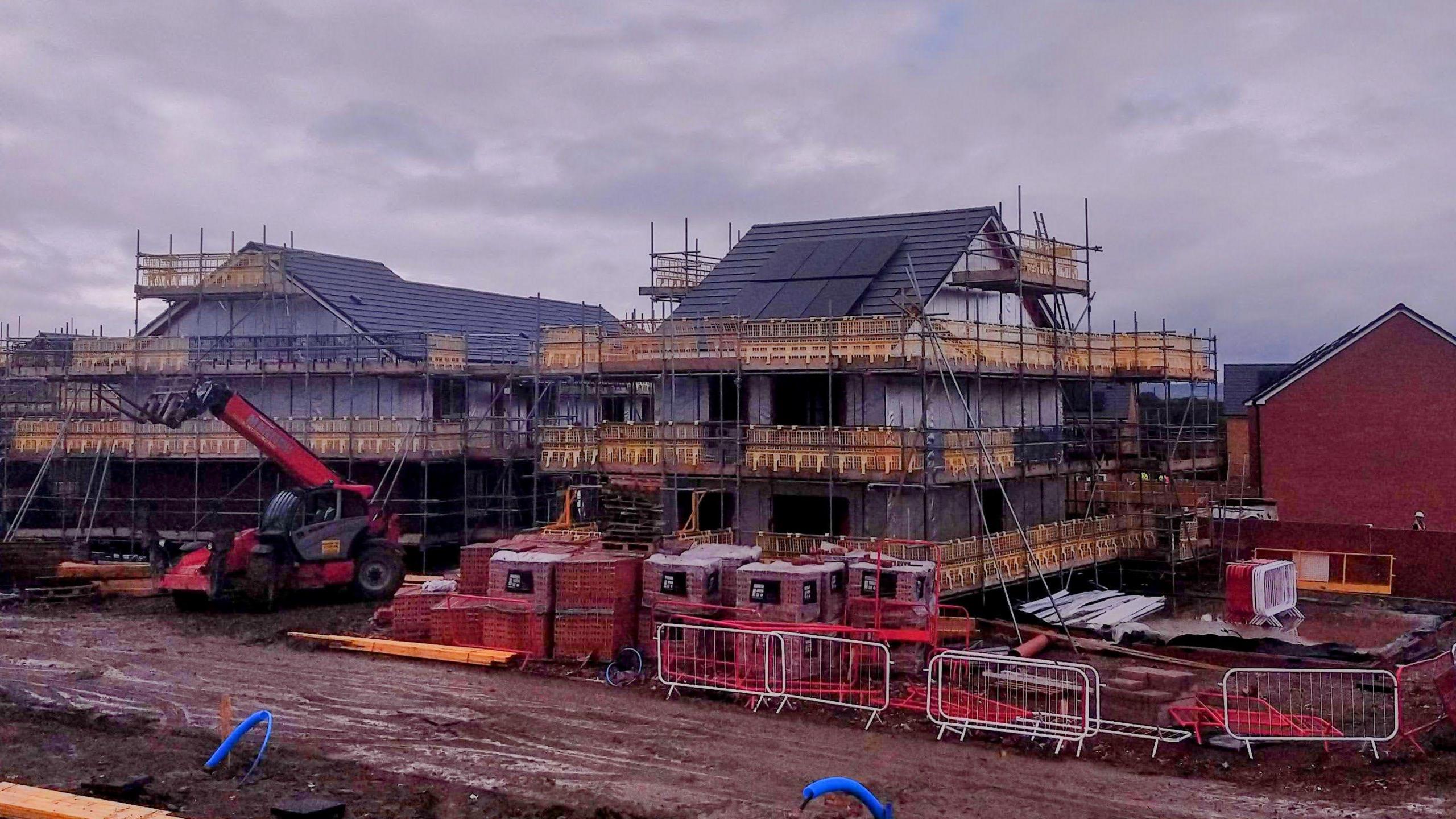 Two houses on a building site covered in scaffolding and surrounded by packaged bricks, surrounded by metal fencing. There is a red digger outside one of the homes and a muddy red track is out the front. Completed houses can be seen in the background and, in the foreground, bright blue pipes are emerging from the mud. It is a very grey day with thick cloud overhead.