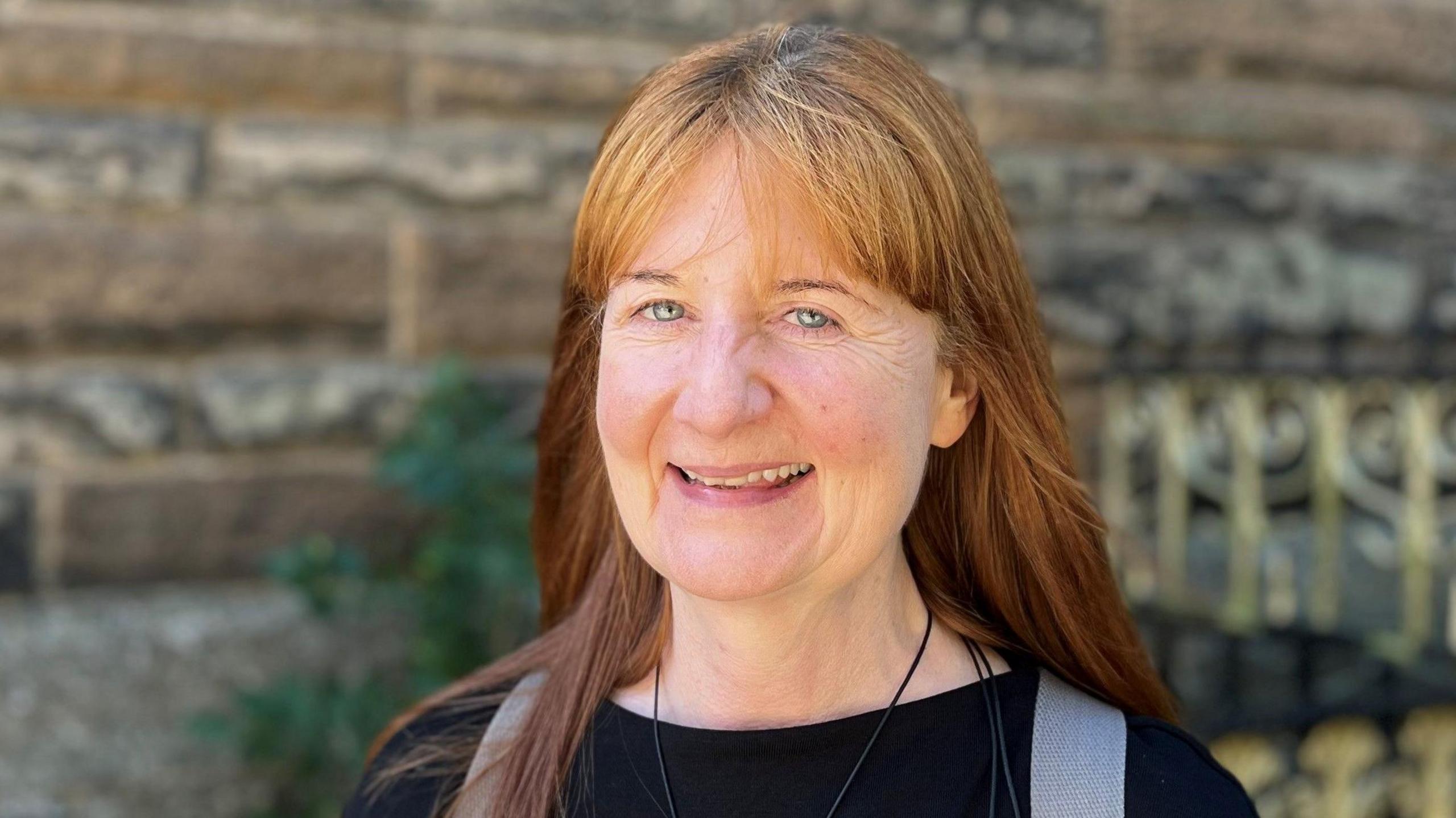Clare Smith smiling at the camera. She has long, lioght brown hair and is wearing a black top. She is standing in front of a sandstone wall.