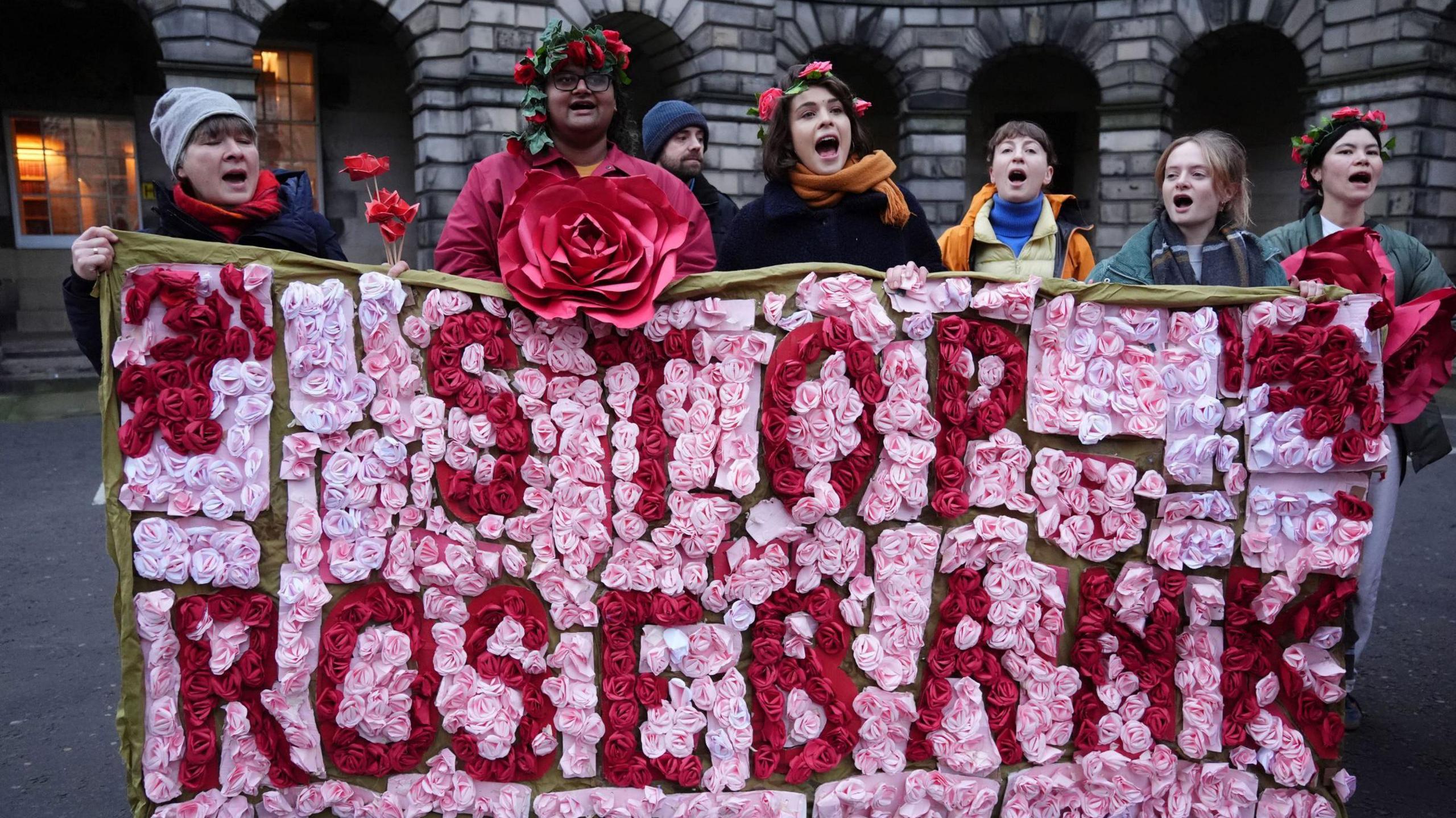 Seven protesters holding a large pink and red floral banner that says STOP ROSEBANK in large letters. The protesters are all shouting or chanting.  