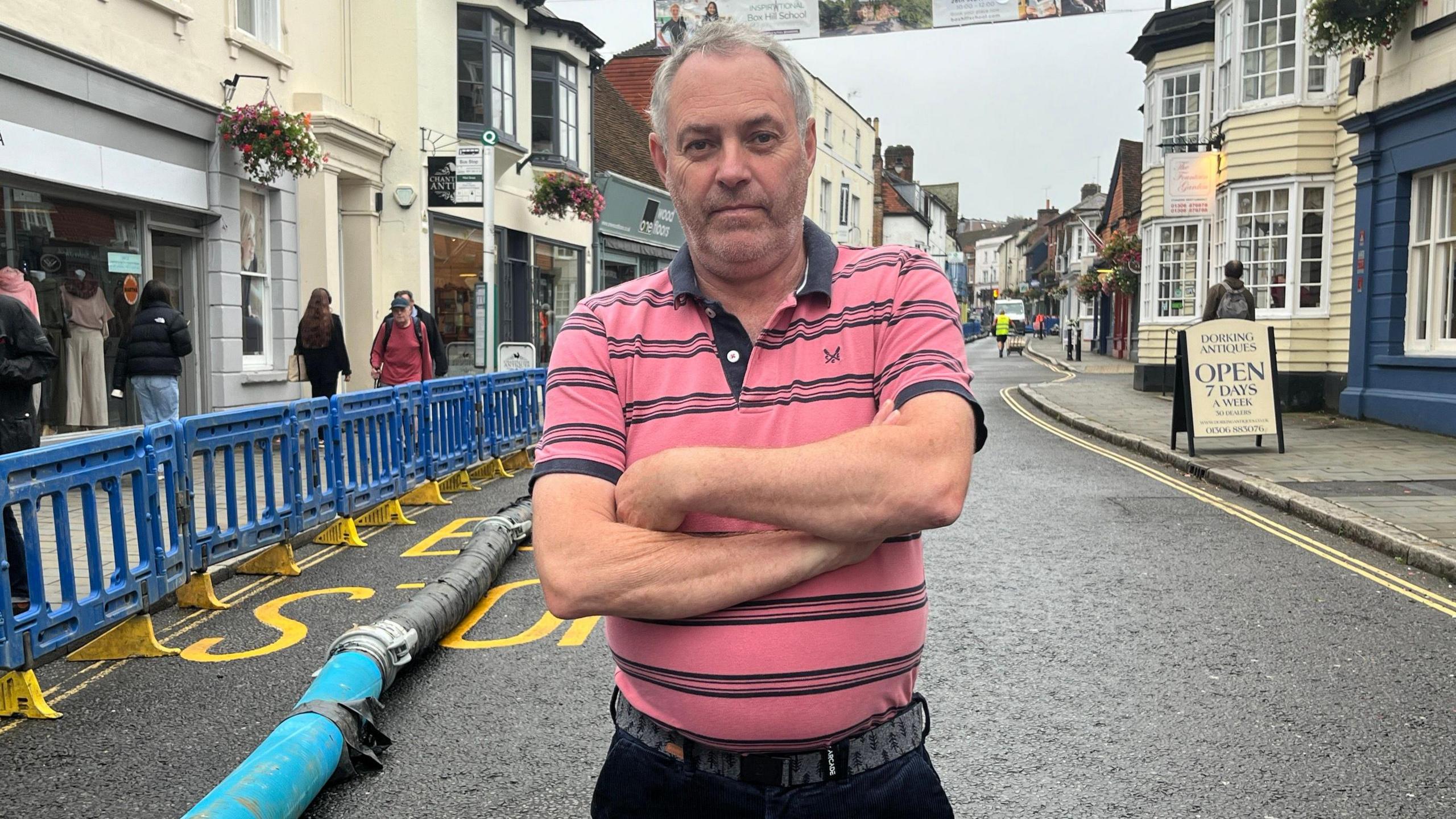 Paul Harman stands in the middle of West Street in a pink and blue striped shirt. On the left are roadworks. A few people are strolling down the street, which has shops and old buildings.