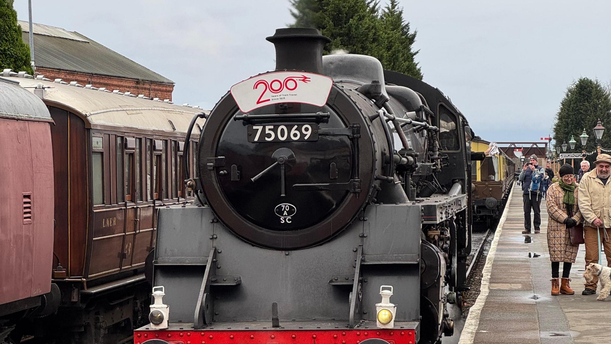 A steam engine stands at a railway station. It has a black front with the numbers "75069" painted on it with a sign above them reading "200". Next to the engine, people mill on the platform in winter clothing.