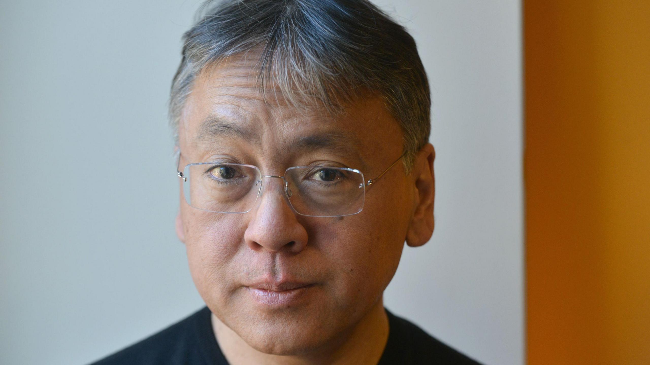 A portrait picture of the writer Kazuo Ishiguro. He has grey hair and is wearing glasses and is staring straight at the camera.