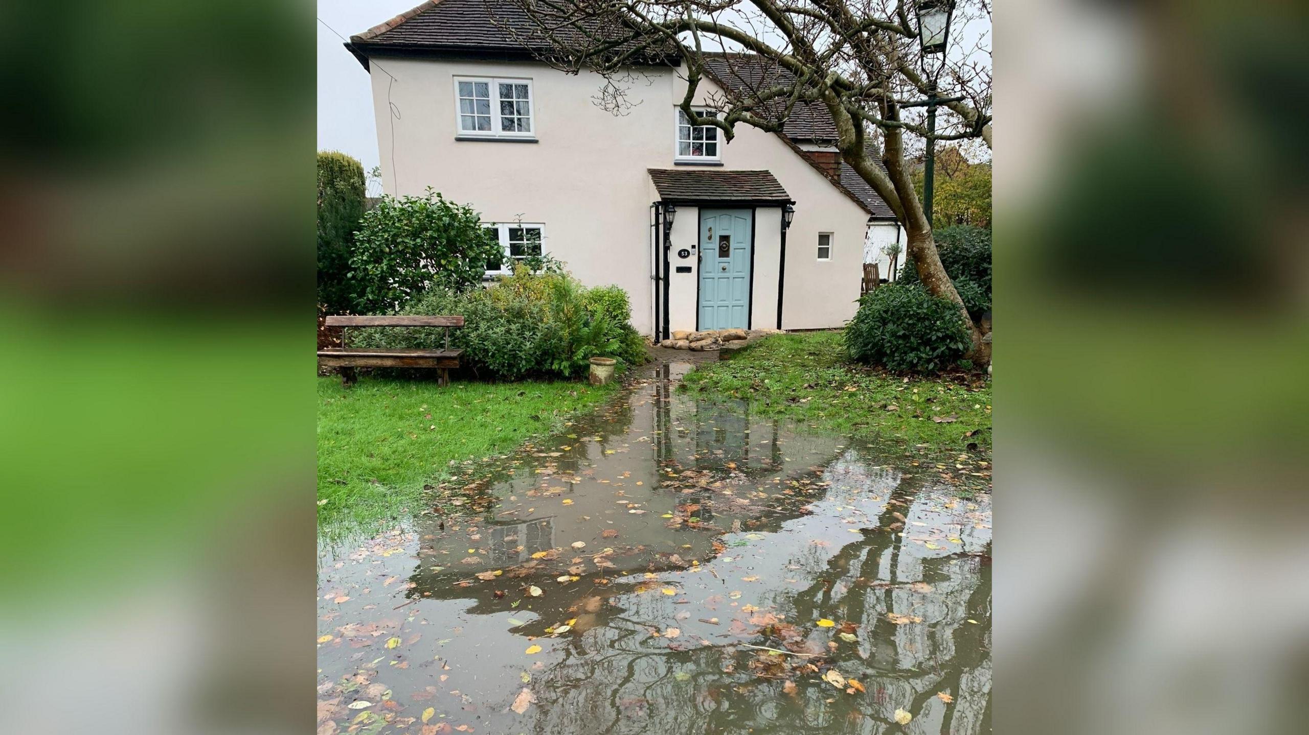 The outside of a two-floor cottage type house with a pale blue front door which has sandbags laid at the bottom of it. There is water in the garden which is covering the lawn and stretching towards the front door but doesn't quite reach it.