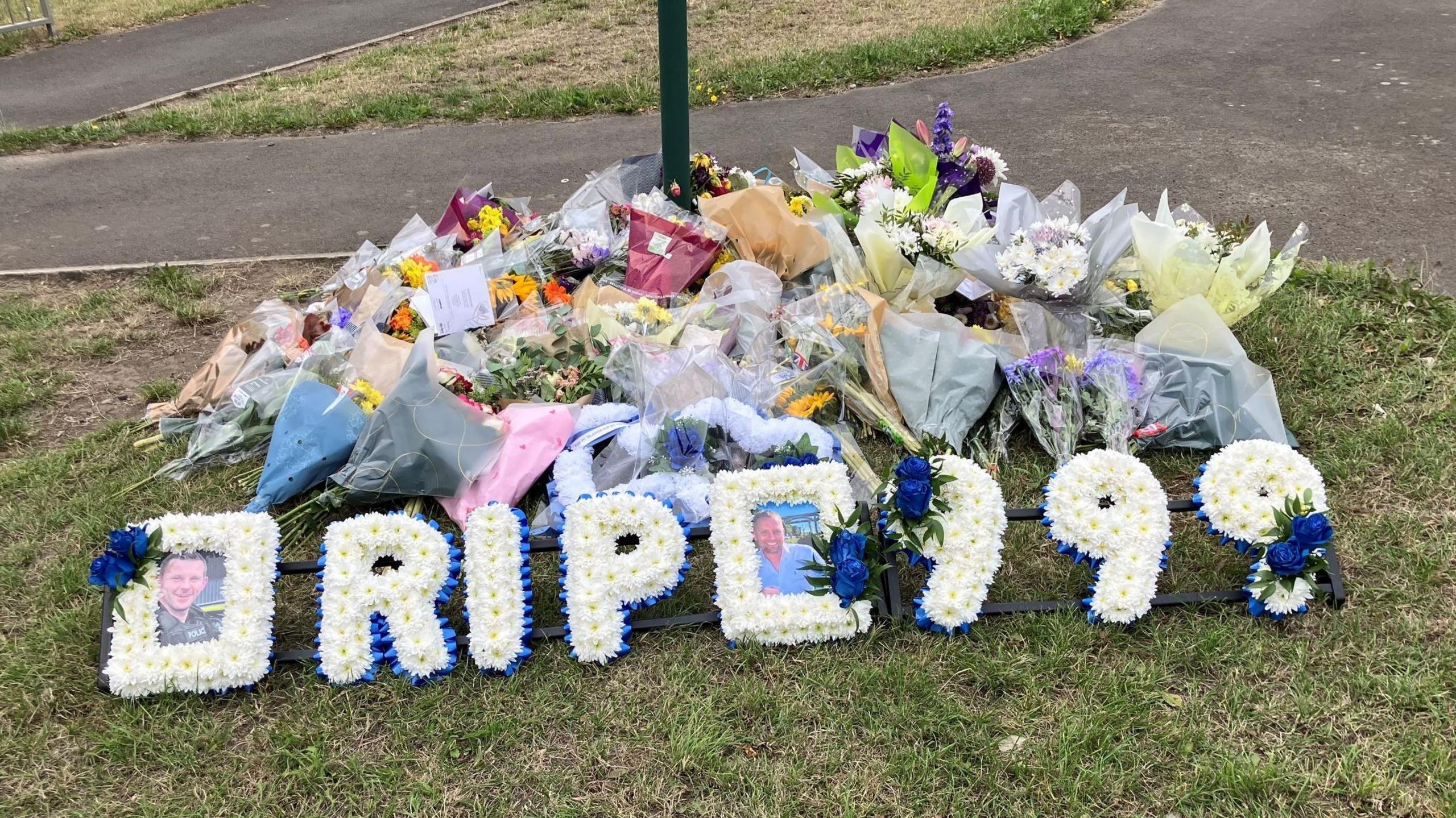 A pile of flowers are placed next to a post in a grassy area next to a pavement. There is a blue and white flower tribute at the top of the pile that spells out: "RIP 999" and has pictures of PC Tom Clough and PC Matthew Blades.